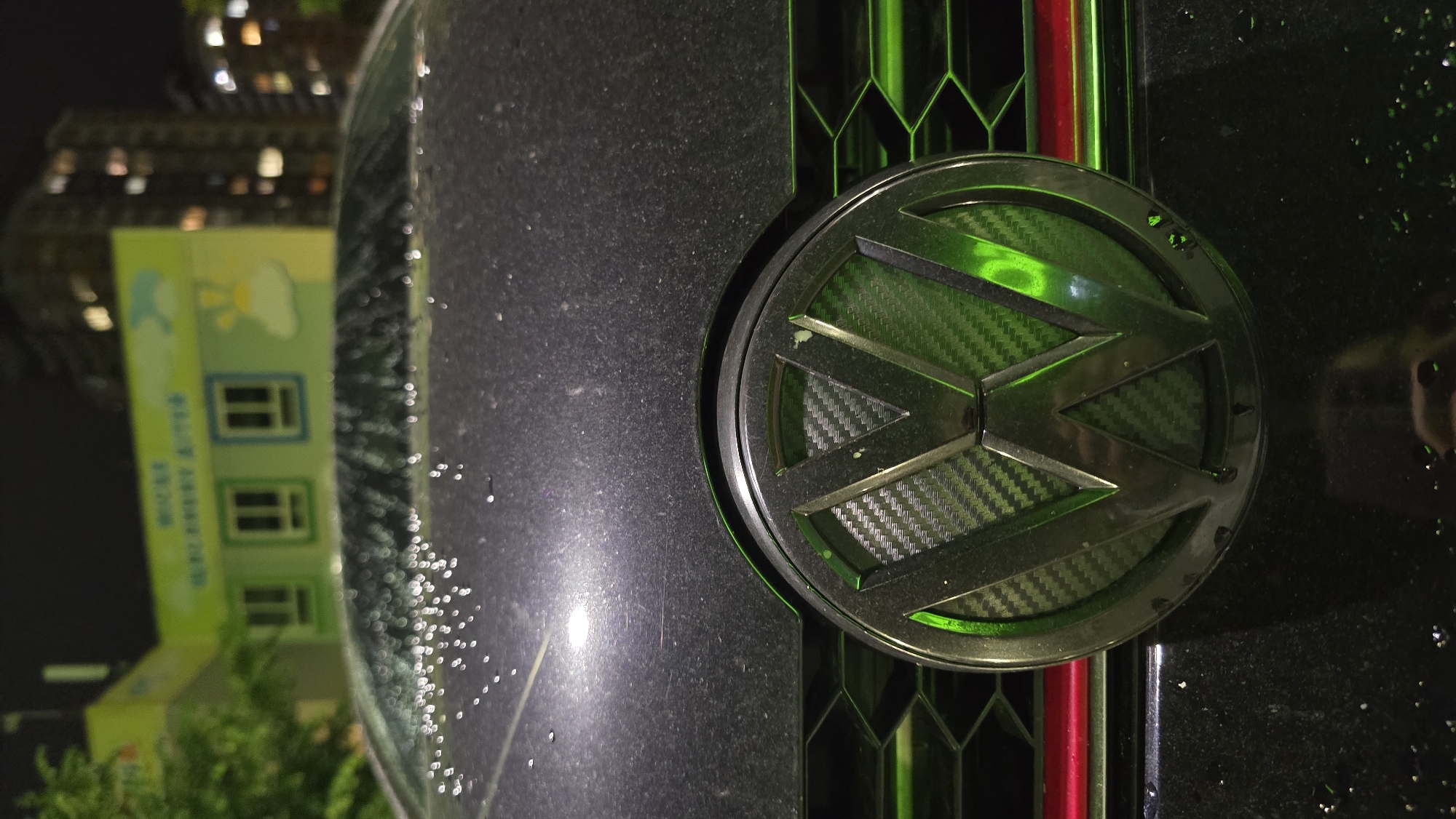The image presents a vertically oriented, close-up view of a dark car at night, focusing primarily on its Volkswagen emblem and part of the hood or grille. The car's surface, particularly on the left, is covered in small water droplets, suggesting it has recently rained or is currently raining. The Volkswagen emblem is dark with a carbon fiber texture visible within the "VW" letters, and it is strikingly illuminated with a vibrant green light. To the right of the emblem, distinct vertical stripes in green and red are visible, likely part of the car's grille design.
In the blurred background, reflected on the car's wet surface or seen through a windshield, is a light green building with multiple windows. At the top of this building, faint yellow and white graphics, possibly depicting a cloud and a sun, are discernible. There also appears to be some blurry text in Chinese characters on the building, but it is unreadable. The overall setting is dark, typical of nighttime in an urban environment, with faint, out-of-focus lights from distant buildings or streetlights adding to the atmospheric glow.