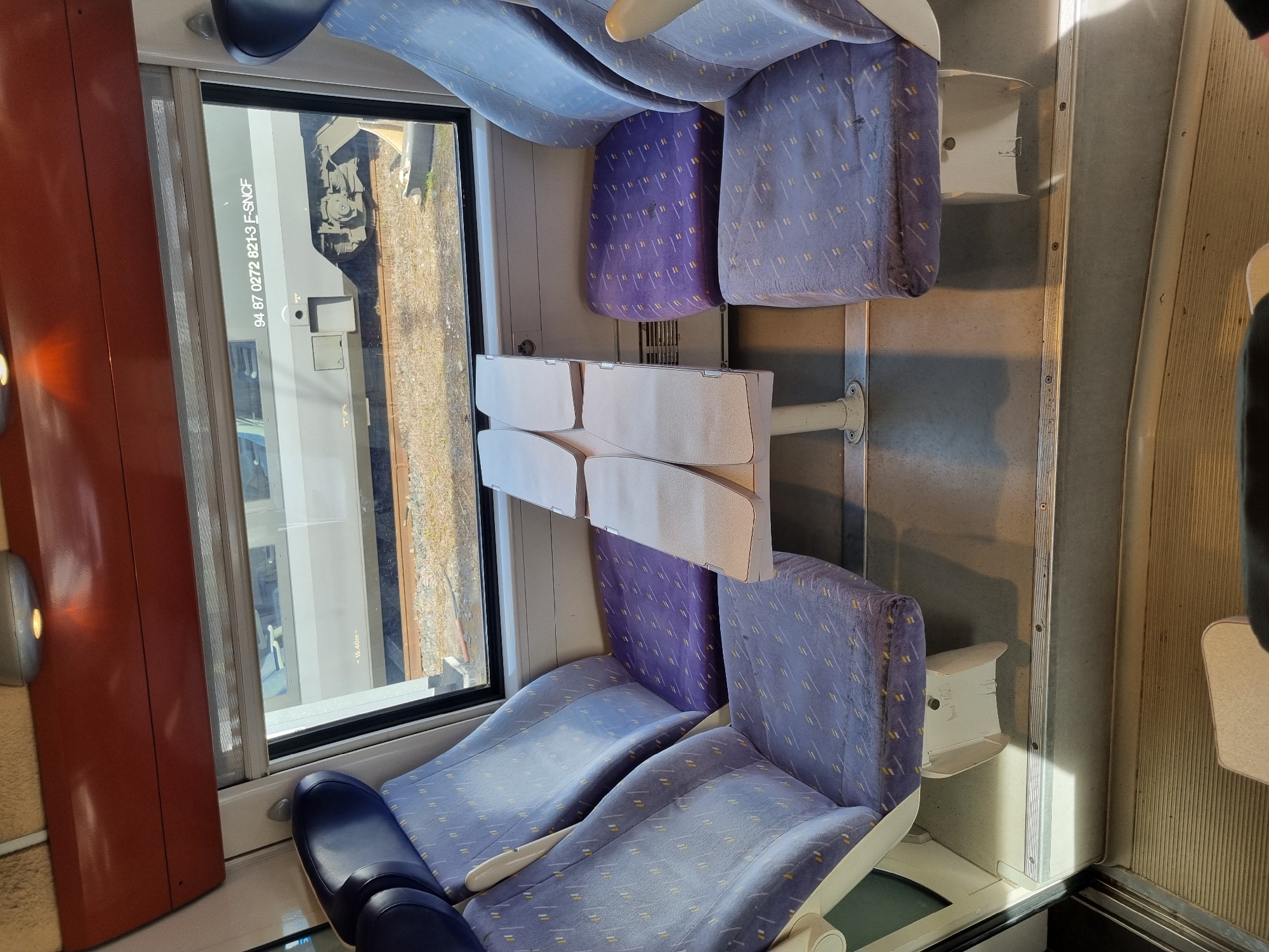 The image shows the interior of a train carriage. Two sets of forward-facing seats are visible, upholstered in a light blue fabric with a patterned design. The seats behind them are folded up in the reverse direction to face the other seats. Each seat has a small, fold-down table attached. A window is on the left, revealing a view of tracks and railway equipment. The number 94 87 0272 821-3 F-SNCF is visible on the side of another train outside the window. Given that the number includes "F-SNCF," which stands for French National Railway Company, the train is located in France. The scene is captured from inside a moving train, providing a partial view of the interior and the exterior through the window. It could be near the city of Forbach, France, considering its location within the French railway system, but this is not certain.