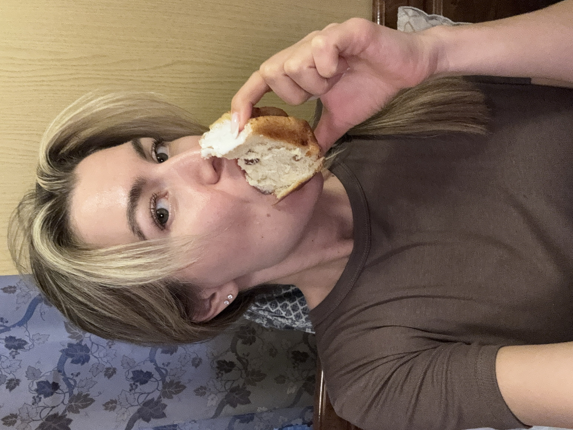 A close-up, rotated portrait shows a woman indoors, holding a piece of bread or a muffin near her mouth. She has light brown hair with blonde highlights, styled around her face. Her eyes are wide open, looking directly at the viewer with an intense or surprised expression. Her lips are slightly parted, and a piece of the white interior of the bread is visible near her nose.
She is wearing a dark brown, long-sleeved top. Her right hand, with neatly manicured nails, holds the bready item. The bread itself appears to be a light brown, crusty exterior with a soft, white interior, possibly containing small dark inclusions like raisins.
The background consists of several elements: a light wooden surface occupies the upper left, suggesting a wall or furniture. On the bottom left, there is a blue and grey patterned fabric, possibly a duvet cover or blanket. To the far right, a darker, possibly wooden, furniture piece is visible, along with a glimpse of a grey and white patterned fabric, likely a pillow or sheet. The overall setting appears to be a casual, indoor environment, possibly a bedroom. No text is visible in the image. The time of day is not discernible from the indoor lighting.