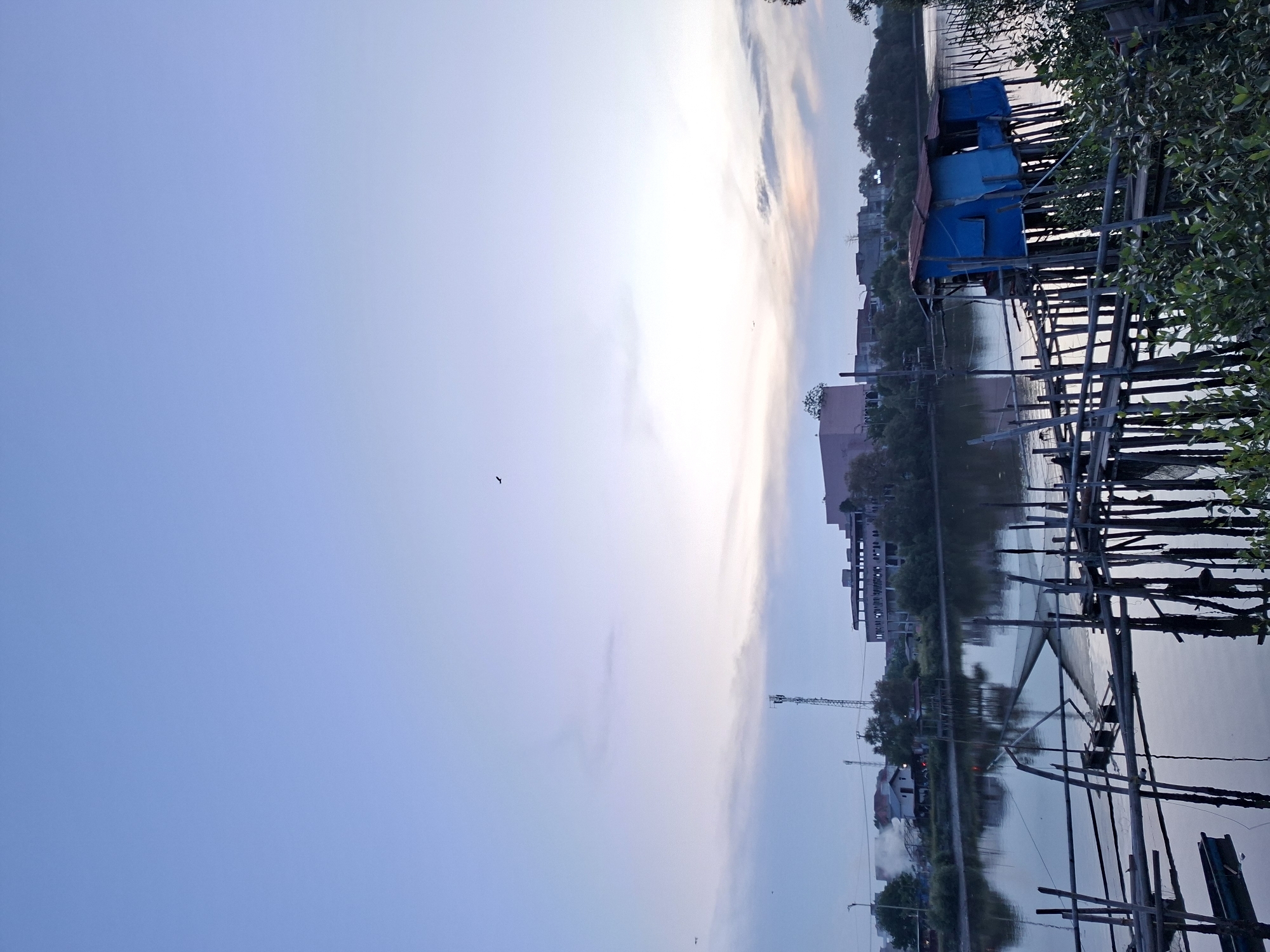 The image depicts a coastal scene in Lhokseumawe, Indonesia, at dusk. The sky is a pale blue with wisps of white and orange clouds near the horizon. In the middle ground are buildings and vegetation. Wooden structures with a small blue building are partially submerged in the water. The foreground features wooden pylons and dense green vegetation. The water reflects the sky and the surrounding scenery, giving it a murky appearance. A bird is flying in the distance.
