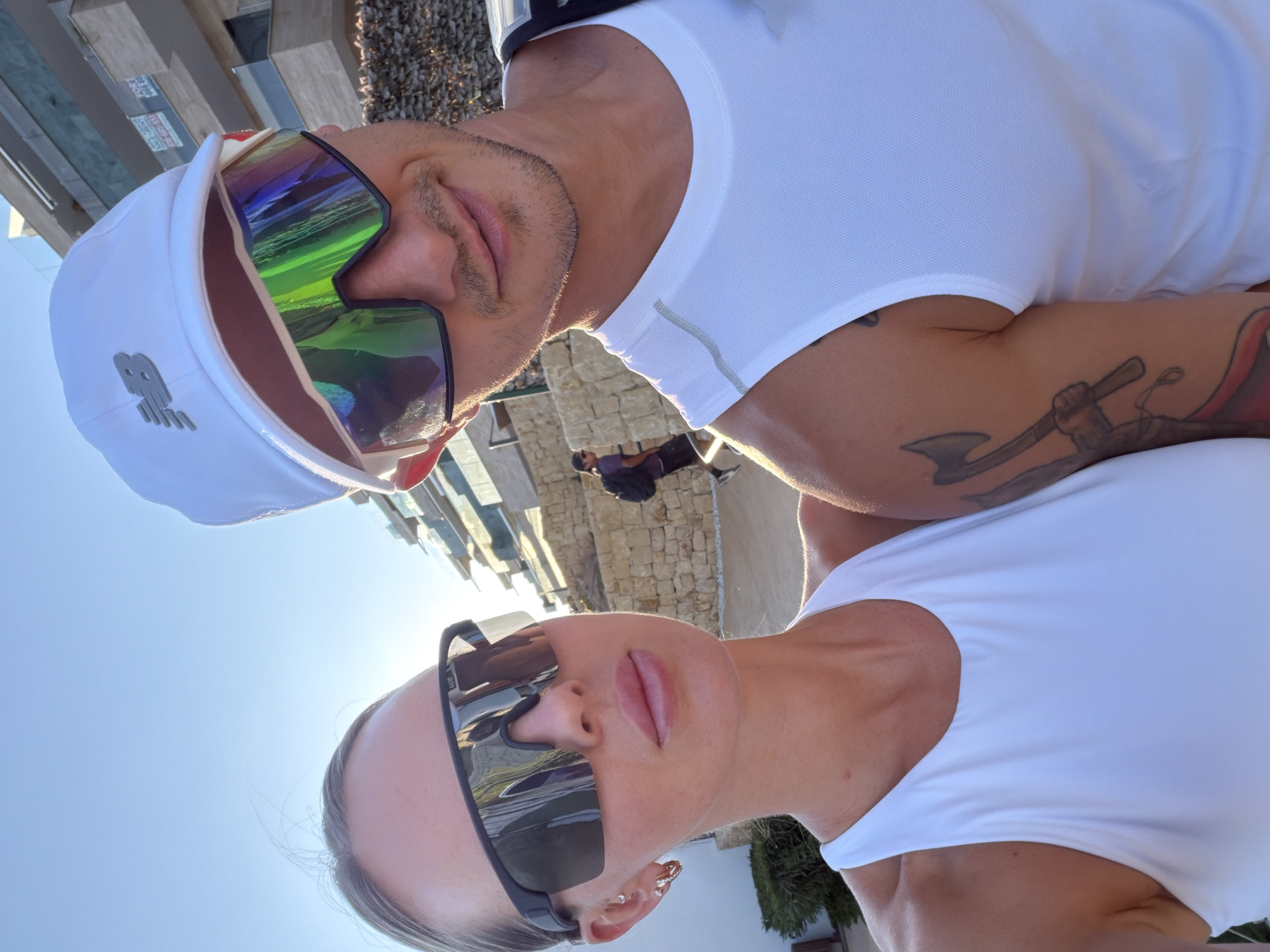 Two individuals, a male and a female, are pictured outdoors in an urban environment on a sunny day. The male, in the foreground right, is wearing a white New Balance baseball cap and iridescent, wraparound sunglasses, and a white sleeveless athletic shirt. He has a light stubble and a tattoo of an axe-wielding figure on his right arm. The female, in the foreground left, is wearing large, dark wraparound sunglasses and a white sleeveless athletic top. She has blonde hair pulled back and is wearing small hoop earrings. Behind them, a building with modern architecture and balconies is visible, along with stone walls and some greenery. A third person, wearing a backpack and dark clothing, is walking in the background. The scene appears to be in Finestrat, Spain.