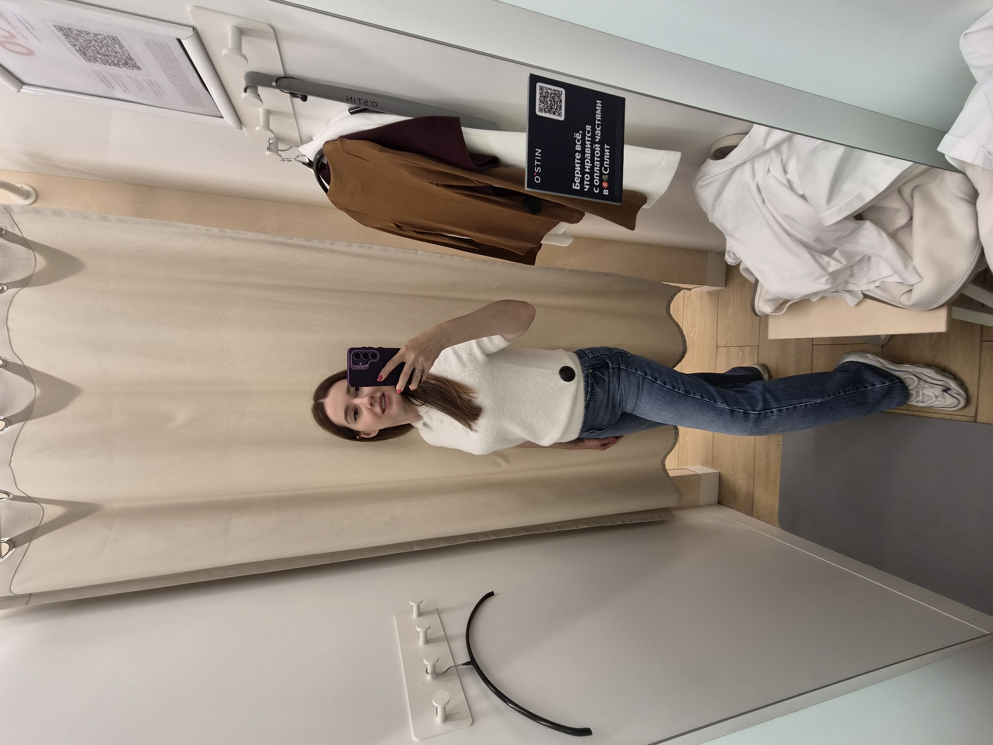 The image shows a young woman with brown hair taking a mirror selfie in a clothing store fitting room. She is wearing a white, fuzzy, collared shirt and blue jeans. She is holding a purple phone in her right hand. In the background, there are beige curtains, a white wall with hooks, and a mirror. A sign in Russian is visible in the mirror, with text that translates to "Take everything you like with installments at Split". There are also several clothing items hanging on a rack and piled on a stool. This scene is consistent with a fitting room in the "O'STIN" clothing store chain, which has locations in Cherepovets, Russia.