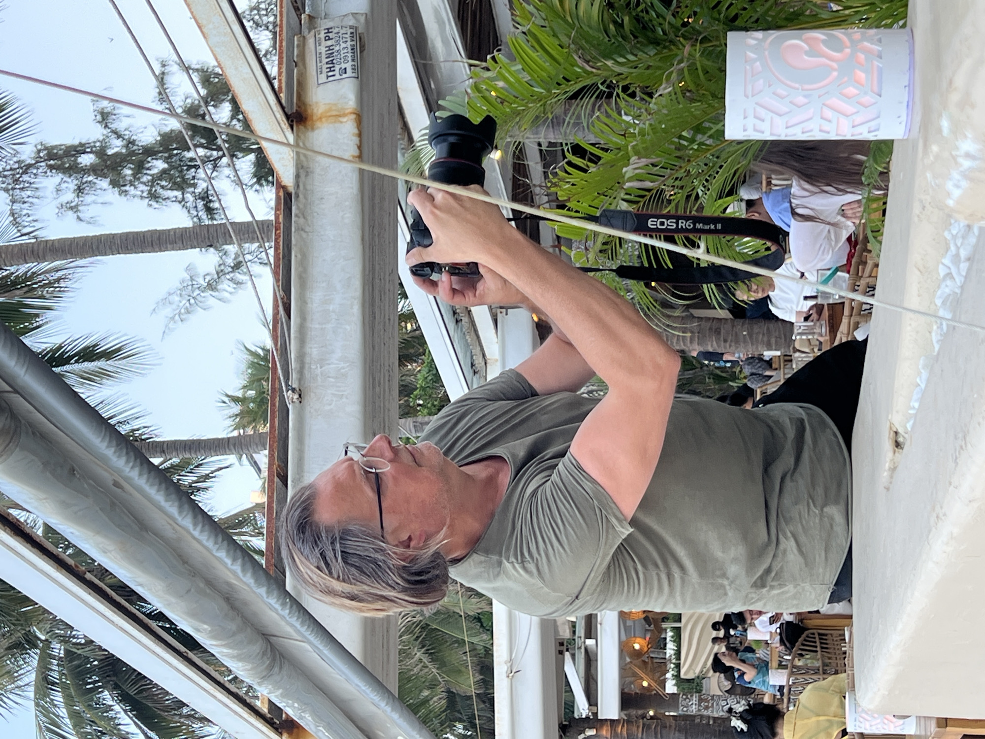 A person is holding a camera, identified by the “EOS R6 Mark II” strap. The person has light-colored hair, glasses, and is wearing a green shirt and black pants. The background includes palm trees and outdoor seating with other people visible in the distance. Text on a structure in the background indicates the location: "183 Hoàng Văn" in Nha Trang, Vietnam. The text on the side reads "MÁI HIÊN - MÀU KE - THANH PH 0258.3824.7 0913.471.7". The scene appears to be at an outdoor dining area or patio with a partial view of the sky.