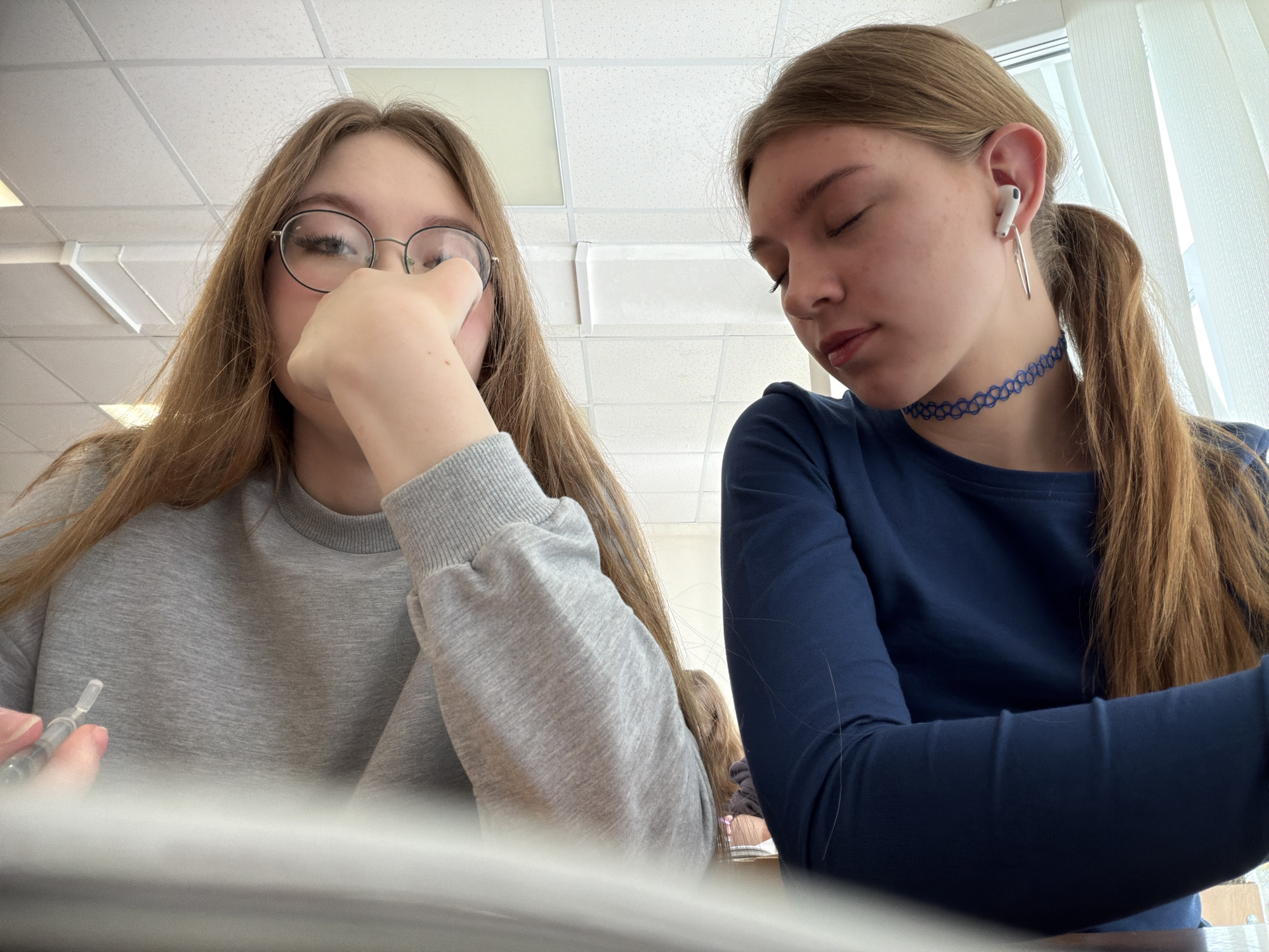 The image depicts two young females seated in an indoor environment, consistent with a classroom or educational facility in Chaikovsky, Russia, indicated by the standard white acoustic ceiling tiles and fluorescent light fixtures overhead.
On the left, a female with long light brown hair and round eyeglasses wears a grey long-sleeved sweatshirt. She holds a clear, cylindrical plastic object, possibly a vial or applicator, in her left hand. Her right hand is raised, with fingers touching the frame of her eyeglasses near her right eye. Her gaze is directed forward.
On the right, a second female with long reddish-brown hair styled in a ponytail wears a navy blue long-sleeved top and a blue woven choker necklace. A white wireless earbud is present in her left ear, alongside a silver-colored dangle earring. Her eyes are closed, and her head is tilted slightly downwards.
A window with a white sheer curtain is partially visible on the far right side of the background. Both individuals are positioned as if seated at desks or tables, with a light-colored surface present in the foreground.
