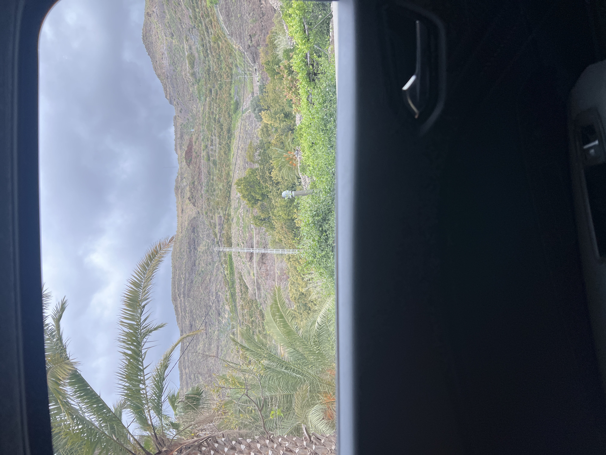The image depicts a landscape seen through a car window, likely taken on a cloudy day in Mogán, Spain. The foreground contains a blurry view of the car's interior including a car vent and door handle. The landscape features a terraced mountain covered in vegetation, including palm trees and other greenery. A tower is visible midway up the mountain. The sky is overcast with grey clouds.