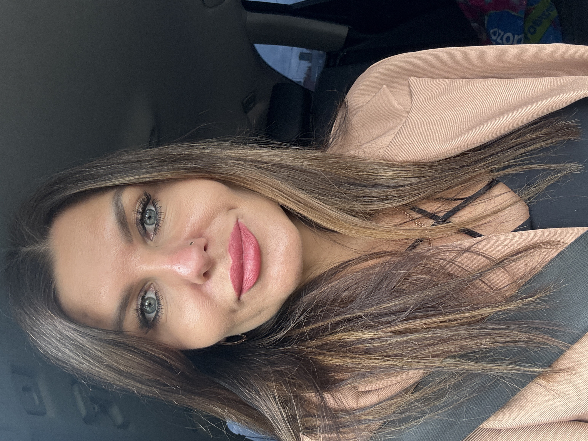 A portrait-oriented photo shows a woman from the chest up, inside what appears to be a car. She has long, dark brown hair with lighter highlights, which frames her face and falls over her shoulders. Her light blue-green eyes are prominent, enhanced with mascara and eyeliner, and her eyebrows are well-defined. She is wearing pink lipstick and has a small silver nose piercing on her right nostril. A delicate gold-colored necklace is visible around her neck.
She is dressed in a dark top with thin straps, partially covered by a light beige or light brown blazer or jacket. A black car seatbelt crosses diagonally over her chest. The background reveals the interior of a vehicle, including dark upholstery and parts of the car's roof and window. In the upper right corner, a blue and white bag is visible with the word "Ozon" printed on it, suggesting a retail or delivery service common in Russia. The woman has a subtle, relaxed smile, looking directly towards the camera. The lighting suggests it is daytime.