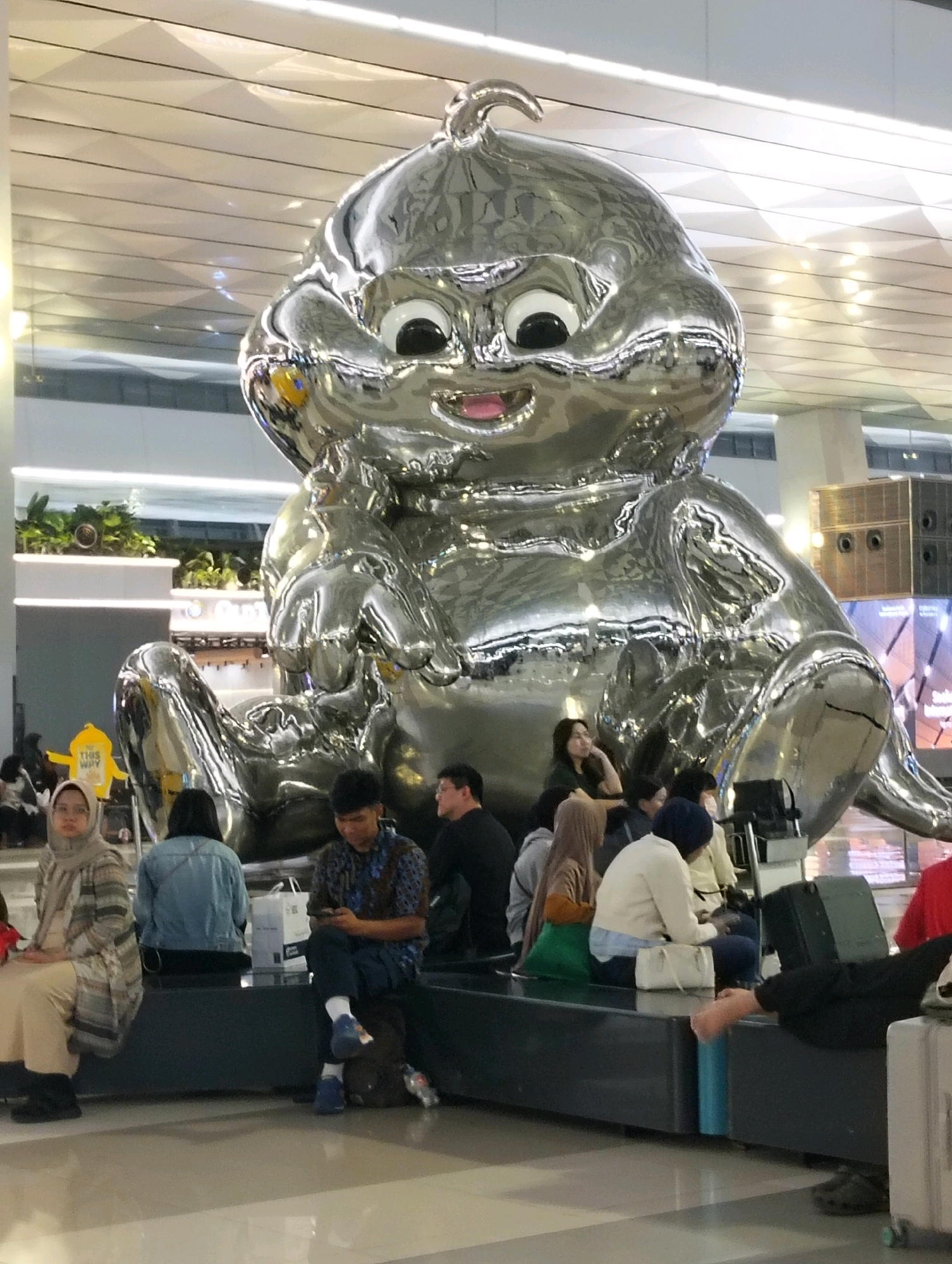 The image captures an indoor scene, likely an airport or public waiting area, with a large, shiny, metallic sculpture of a cartoonish character as the central focal point. The sculpture is silver and has a reflective surface, showing distortions of the surroundings. It is seated with its legs bent and arms resting on its lap.
Several people are seated on benches in front of the sculpture, appearing to be waiting. From left to right, a woman wearing a hijab and a patterned scarf is seated with her legs crossed. Next to her is another woman in a denim jacket. A young man in a patterned shirt is looking at his phone, with his legs crossed. Further to the right, a woman in a beige cardigan and a blue headscarf is seated, and next to her is another woman in a white sweater and blue pants. Some people are looking at their phones, while others are looking around. The environment features a polished floor, a high ceiling with geometric patterns, and some greenery on elevated shelves in the background. There are also luggage items visible, including suitcases and bags. A yellow sign with "THIS WAY" written on it is partially visible in the background. The overall impression is one of a public space with a prominent art installation.