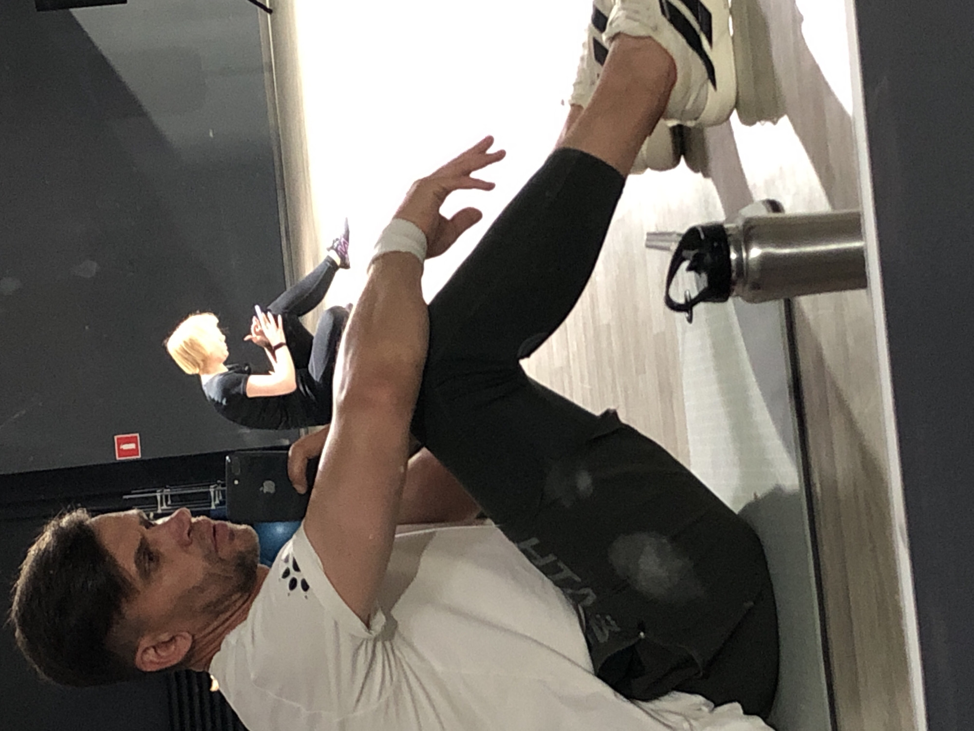 The image depicts an indoor setting, likely a gym, with two individuals present. The primary subject, a male, is in the foreground, seated on a yoga mat, facing left with his body angled towards the camera. He is wearing a white t-shirt and dark athletic leggings, with white sneakers. His left arm is resting on his left leg, and his right hand is extended with fingers splayed. A water bottle is visible on the floor to his right. In the background, a second individual, a female with blonde hair, is seated on the floor, looking down at a phone. The environment appears to be a studio with a dark wall and a light-colored floor. The lighting suggests it is daytime, possibly with artificial light contributing to the illumination. There is no specific visual information in the image that indicates the location is Saint Petersburg, Russia.