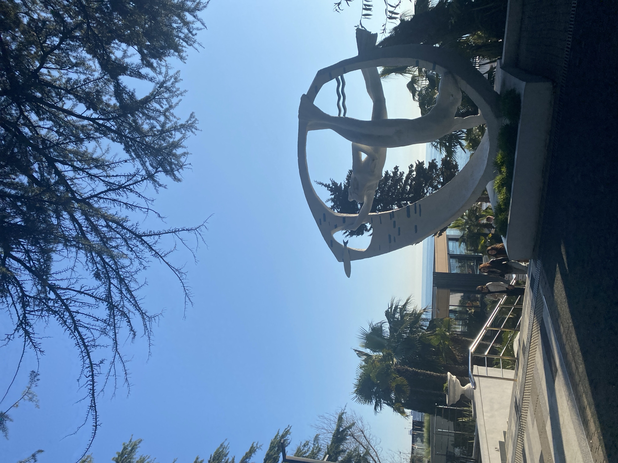 The image shows a low-angle view of a sculpture and surrounding environment, possibly taken in Sochi, Russia. The sculpture, in the center of the frame, is a white, circular structure with stylized human figures inside. The environment includes trees, including some palm trees, suggesting a coastal location. There is a building visible in the background, as well as a set of stairs with metal railings, and a group of people. The sky is blue.