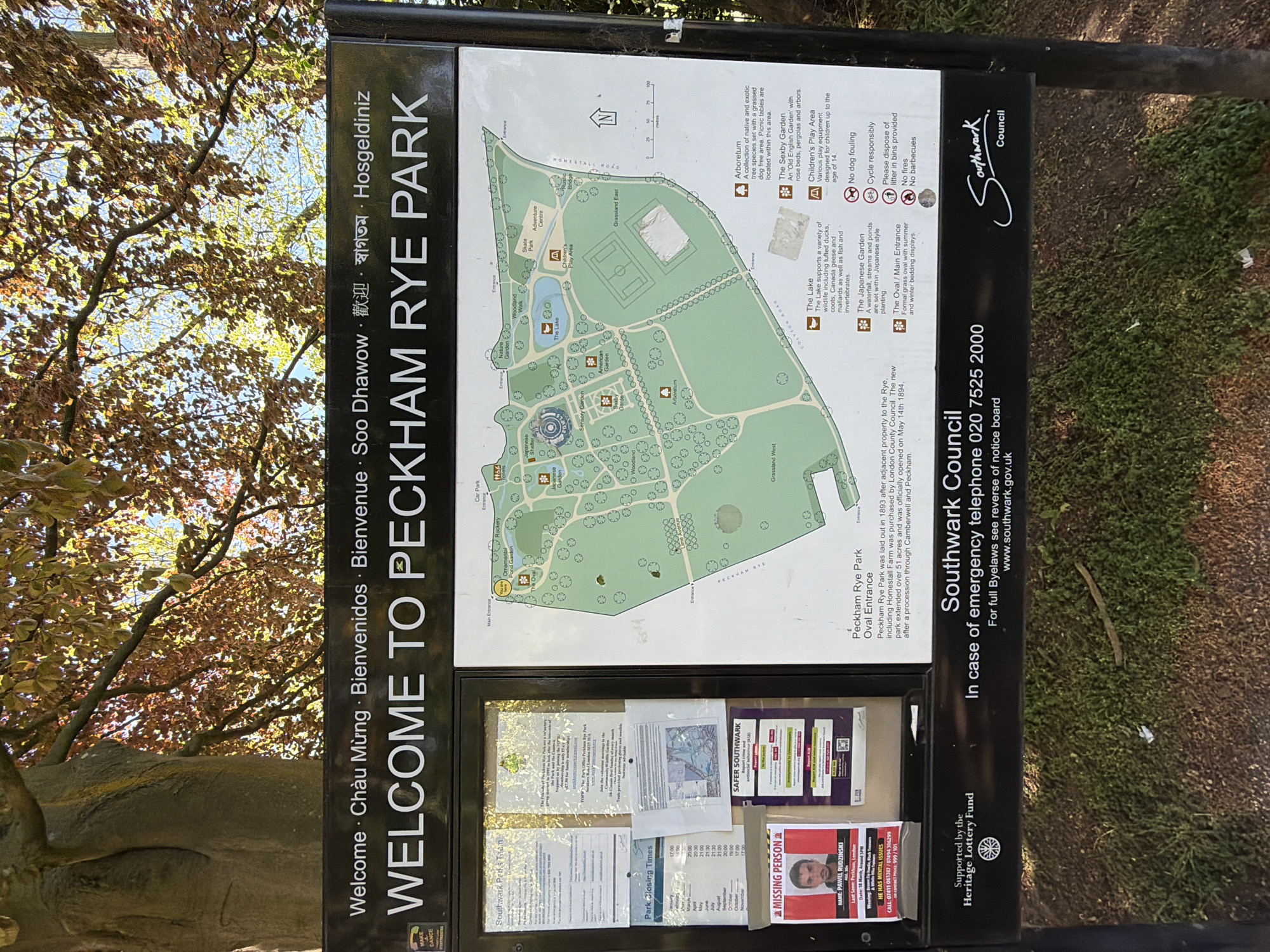 This is an image of a park sign located in Peckham Rye Park in London, United Kingdom. The sign provides a welcome message in multiple languages and a map of the park's layout, including the location of various features such as the Arboretum, Sexby Garden, Children's Play Area, and the Japanese Garden. The sign also includes contact information for Southwark Council and emergency services, along with park rules and regulations, such as no dog fouling, responsible cycling, and proper litter disposal. A notice board at the bottom of the sign displays various community notices, including information about park support organizations, park closing times, a Safer Southwark notice, and a missing person poster. The area surrounding the sign includes a tree and grass.