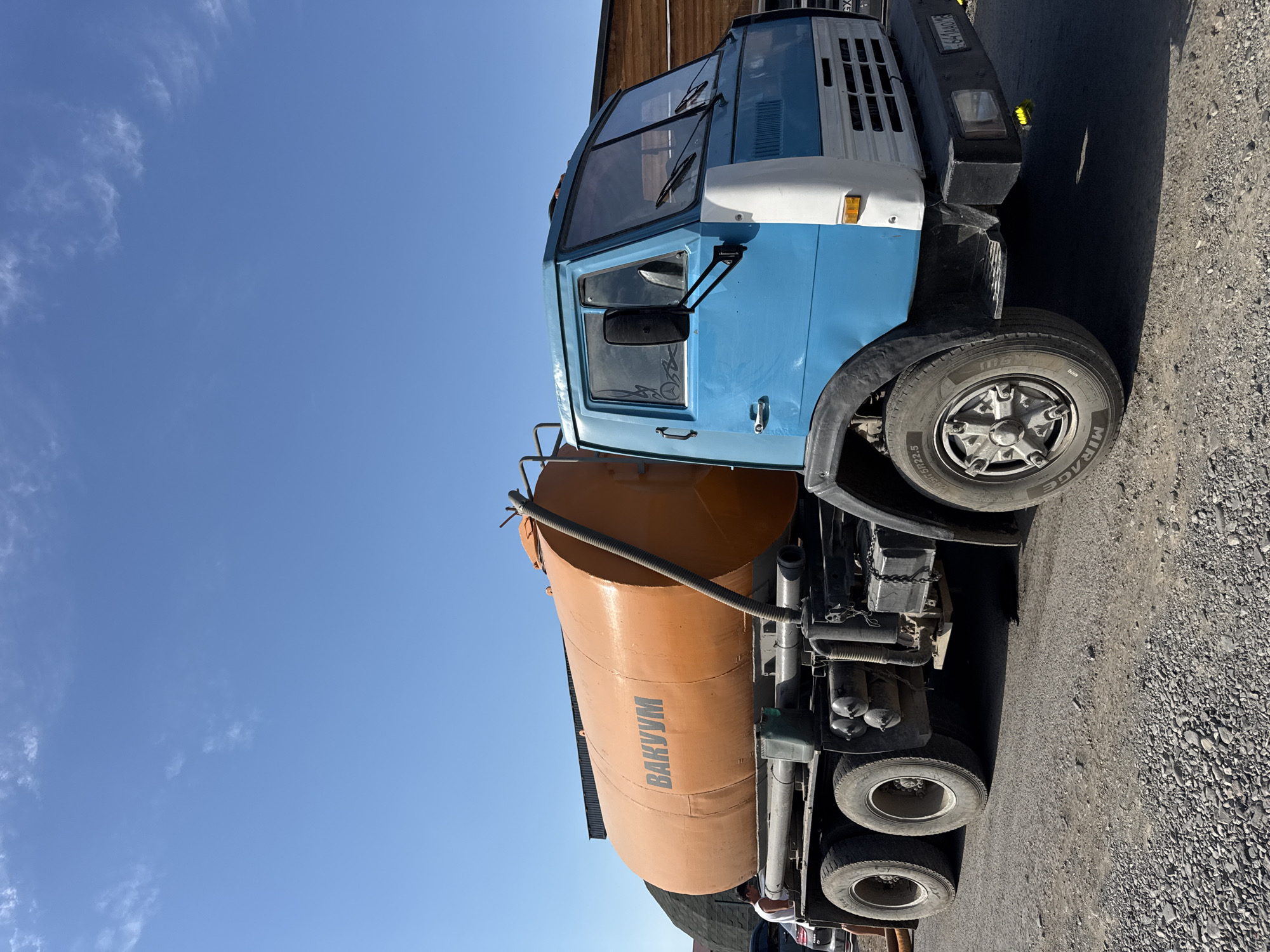 An eye-level, medium shot captures a blue and white KAMAZ truck with an orange vacuum tank positioned on a gravel surface under a clear blue sky. The truck, which is the primary subject, is facing left with its front tire visible. The orange tank, mounted on the truck, displays the Cyrillic word "BAKYM" in dark letters. A partially visible person, wearing a white tank top, stands to the left of the truck. In the background, a wooden structure and a dark, conical pile of material are present. The ground is covered in coarse gravel.