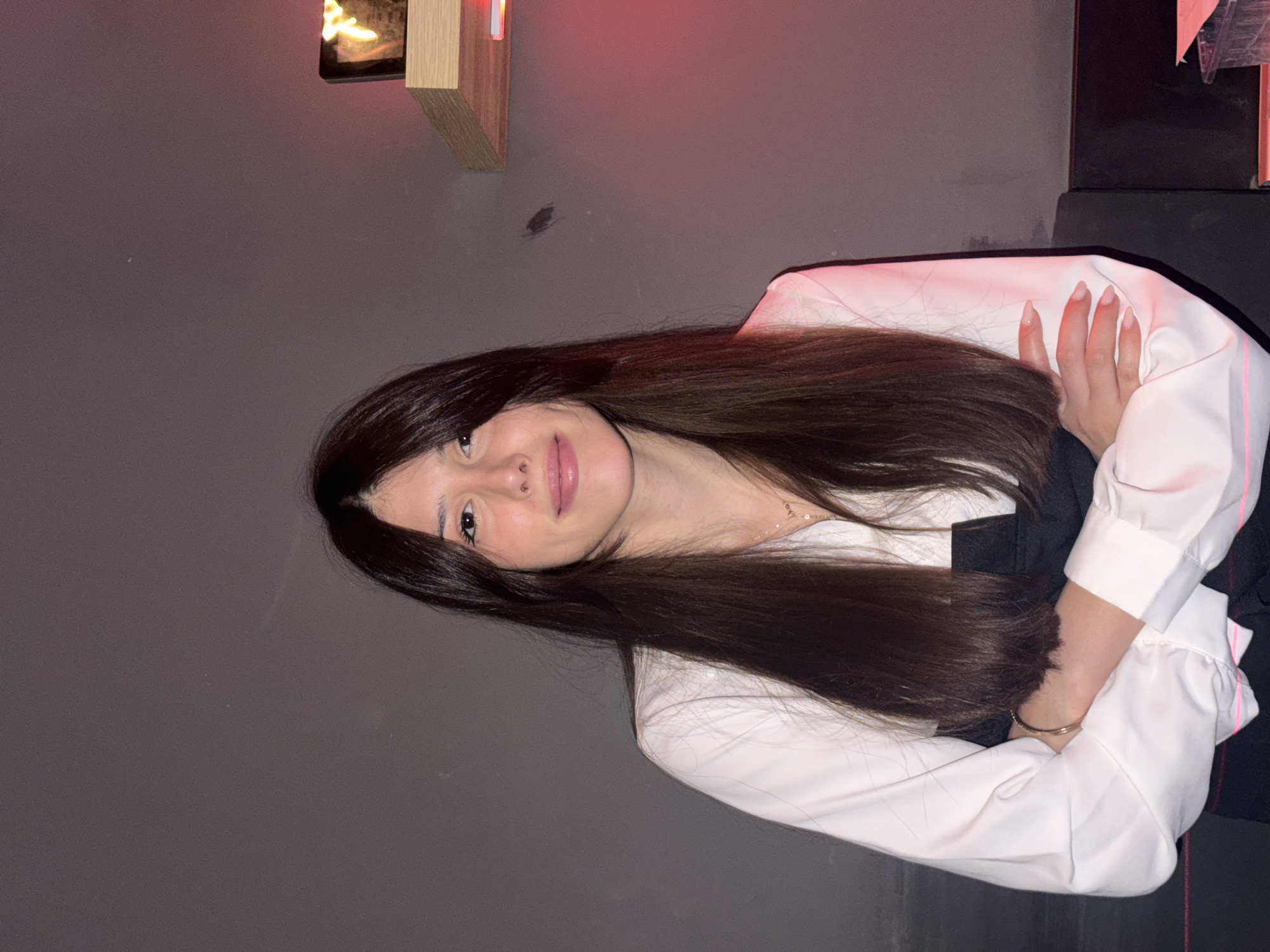 A young woman with long, dark hair and a white blouse with a black top is posing for a photo against a gray wall. Her arms are crossed, and she is looking at the camera with a slight smile. A shelf with a red light is visible in the background to the right of the woman. The environment appears to be indoors.