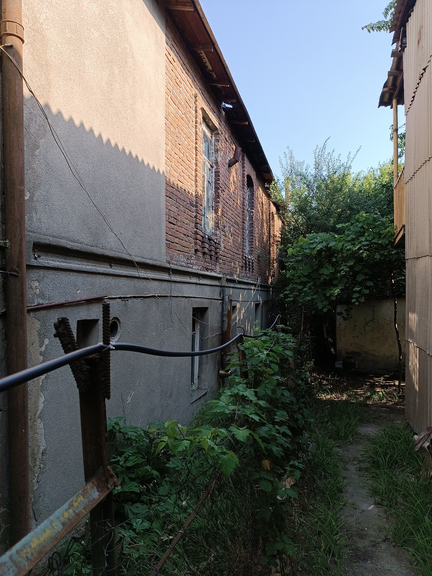 This image depicts a narrow alleyway between two buildings in the city of Khashuri, Georgia. The building on the left is constructed of brick with some areas of stucco and features multiple windows. A rusty pipe runs vertically along its side, and wires are strung across its facade. Overgrown green vegetation, including bushes and tall grass, encroaches on the alleyway, partially obscuring a small, weathered structure visible in the background. The building on the right is clad in corrugated metal siding. The alleyway itself appears to be a dirt path with grass growing along its edges. The sky overhead is clear and blue.