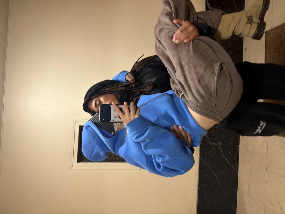 A mirrored selfie features a person wearing a blue hooded sweatshirt and black head covering with dreadlocks. They hold a phone with a patterned case. Behind them stands another person in brown sweatpants, a black jacket, and light brown boots. The background includes a plain wall with a window and a tiled floor with black and white stripes.