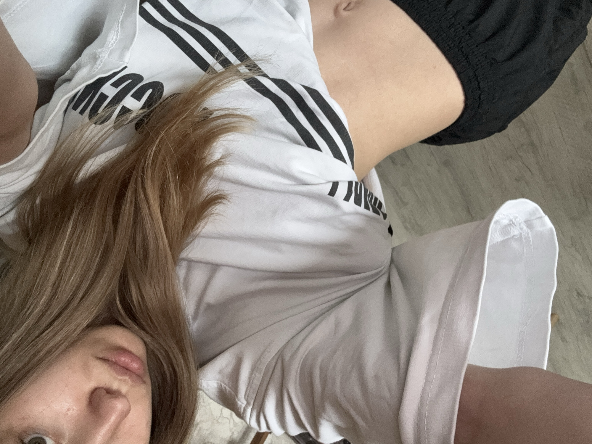A woman with long brown hair is partially visible in the lower left corner of the frame. She is wearing a white t-shirt with black stripes on the sleeves. The t-shirt also has some lettering on it, but it is difficult to read. She is also wearing black shorts. The background appears to be a wooden floor. There is no indication of the location being in Noviy, Russia.