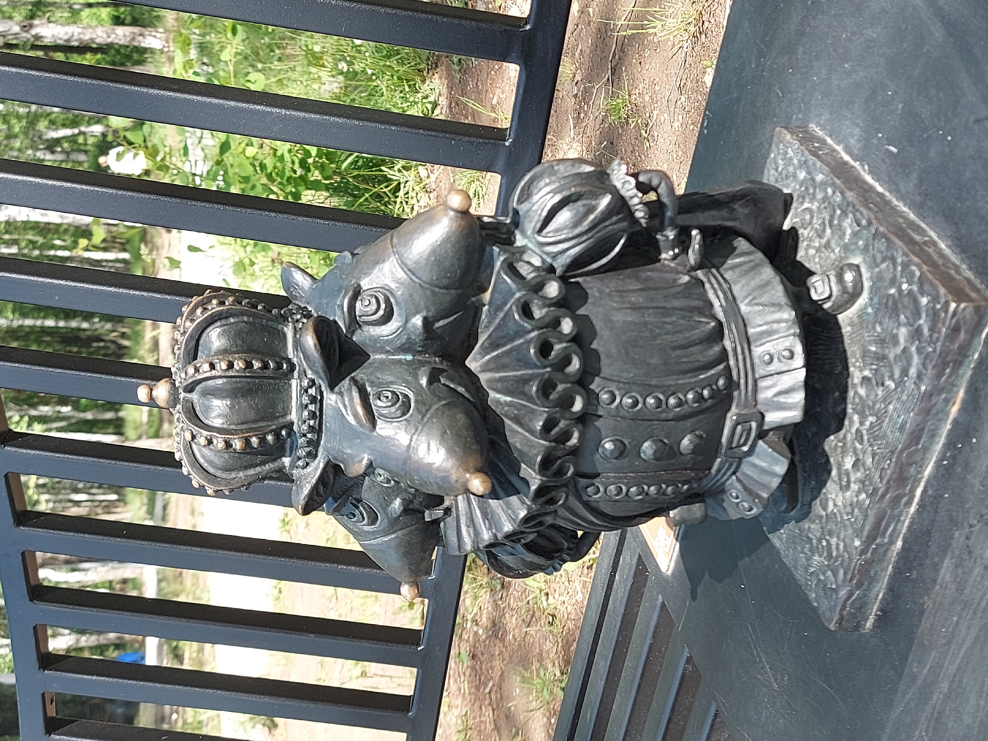 The image shows a bronze sculpture in the city of Chaikovsky, Russia. The sculpture depicts two anthropomorphic rats dressed in regal attire. One rat wears a crown and the other appears to be wearing a ruffled collar and vest. The sculpture is mounted on a square bronze base and is situated next to a black metal fence. The background shows foliage and glimpses of other trees. The scene is outdoors, likely in a park or public space.