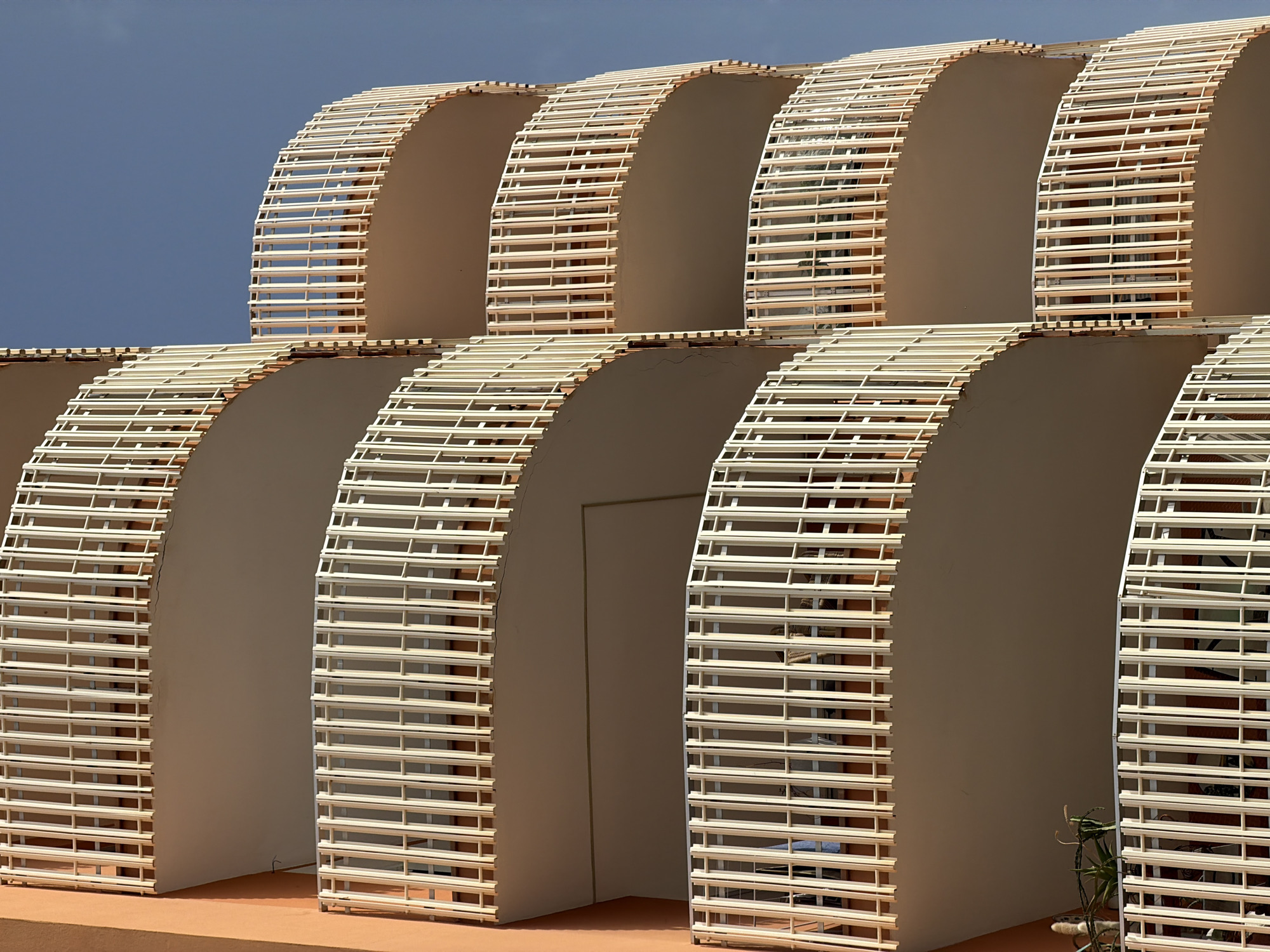 The image displays a segment of an apartment building with multiple curved balconies in Magaluf, Spain. The building's facade is painted in a peachy-orange hue. Each balcony features a white railing composed of vertical slats with horizontal crossbars. The balconies are tiered, with the lower ones partially obscuring the ones above. The sky in the background is a clear, light blue. There are no people visible in the image. The overall scene depicts architectural elements of a residential building under a bright sky.
