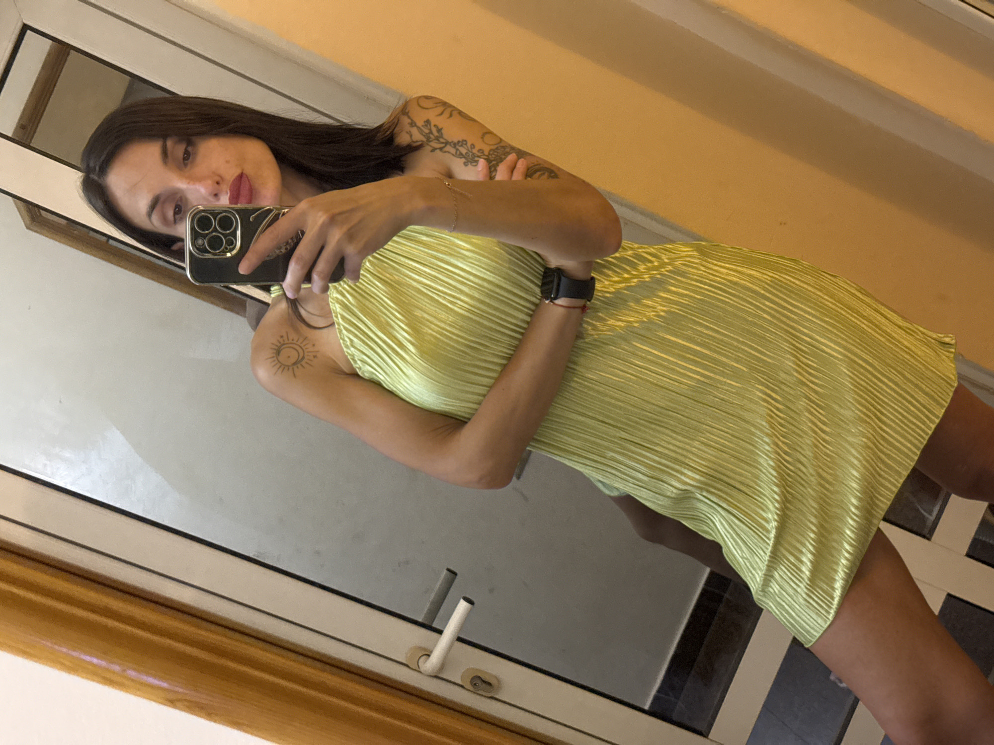 A woman is taking a mirror selfie in what appears to be a room with a doorway and a light-colored wall. She is wearing a light green, sleeveless, pleated dress. Her left arm is raised, holding a black smartphone to take the picture. She has dark hair, light skin, and visible tattoos on her left shoulder and arm. She is wearing a dark watch on her left wrist and a thin bracelet on her right wrist. The background shows a closed door with a white handle and a lock, a large mirror, and a wall with warm lighting. There is no visible indication of the location being in the city of Pleven, Bulgaria.