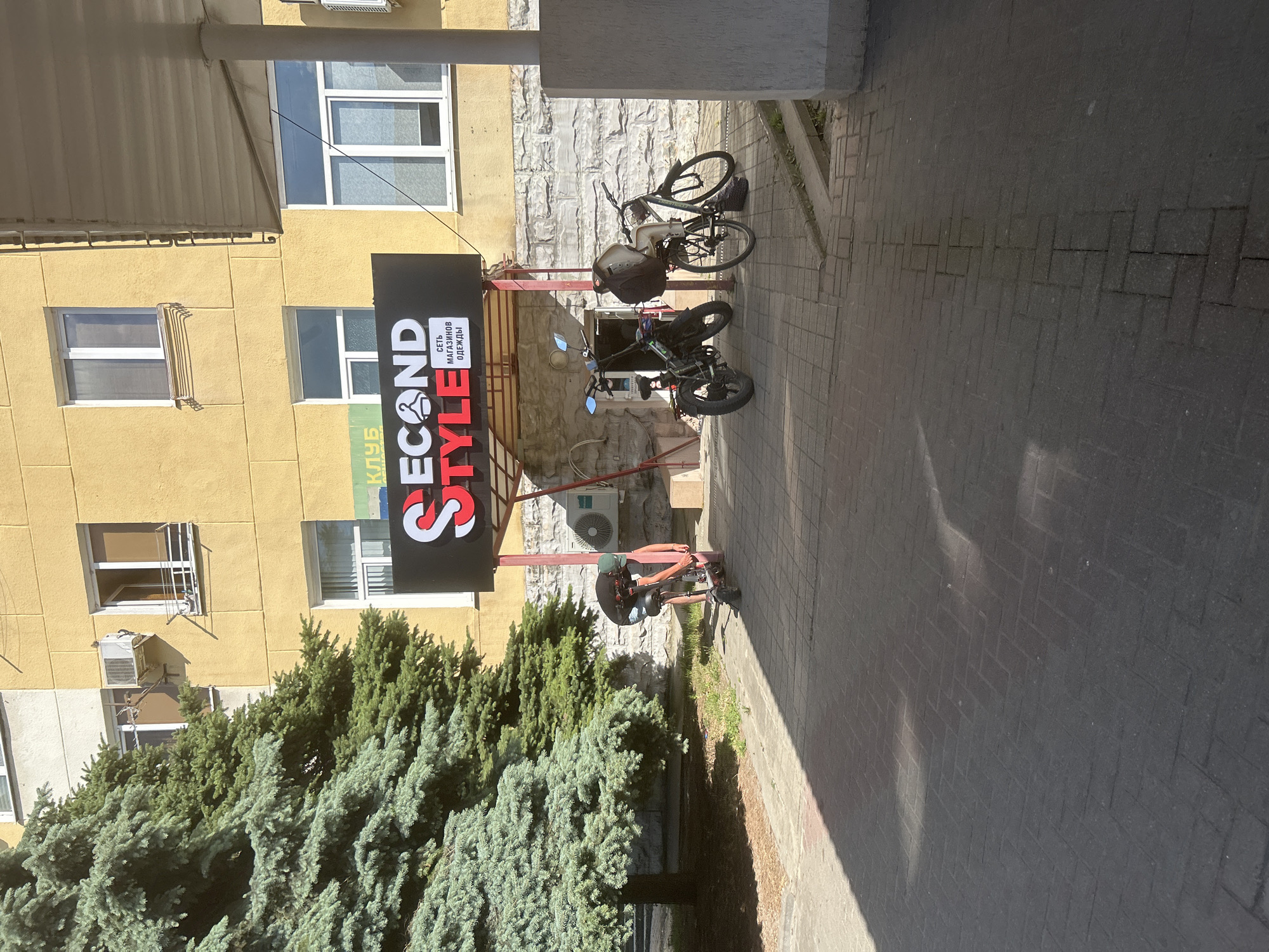 In the city of Gelendzhik, Russia, an outdoor scene captures the facade of a yellow building. Prominently displayed on the building is a black sign with red and white lettering that reads "SECOND STYLE," featuring a hanger icon. Beneath this main sign, a smaller white sign states "СЕТЬ МАГАЗИНОВ ОДЕЖДЫ" (Clothing Stores Network). To the left of the sign, a large green evergreen tree stands tall, casting shadows on the paved walkway. In front of the building's entrance, an individual wearing a dark t-shirt, shorts, and a green cap is interacting with a red electric scooter, seemingly preparing to ride it. Further to the right, two parked vehicles are visible: a black electric bicycle with a large bag on the back and a blue bicycle with a basket. The ground is paved with interlocking gray bricks, and a concrete pillar supports an overhead awning. The overall environment suggests a commercial area on a sunny day.