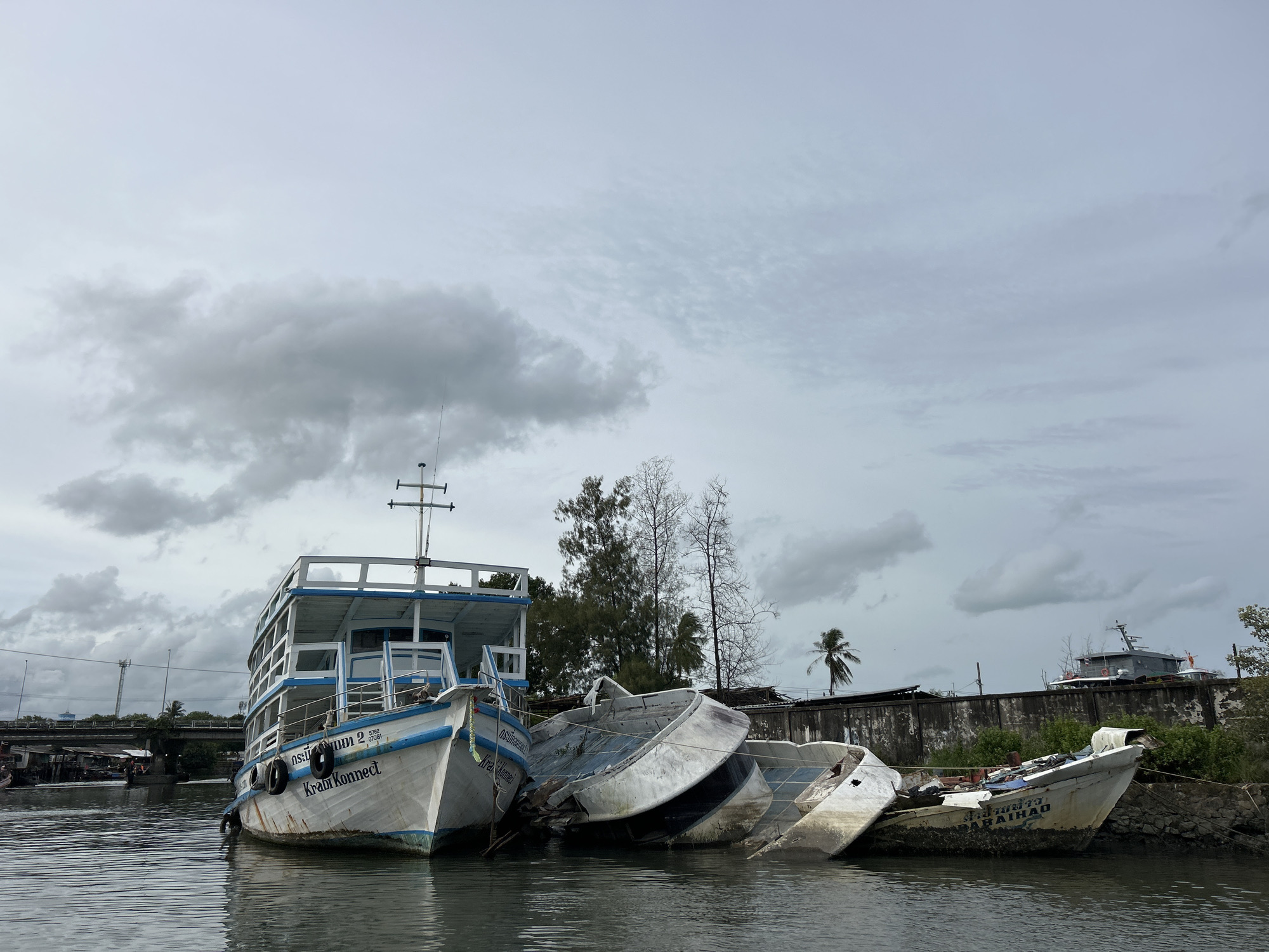 In Ratsada, Thailand, a tranquil waterway is marred by the sight of several abandoned, damaged boats. The largest, a white and blue vessel named "KraorKonnect," sits upright, listing slightly. Alongside it, three other boats lie capsized and broken, their hulls resting against each other in a chaotic heap. The water reflects the overcast sky, where heavy grey clouds gather, hinting at impending rain. Lush green vegetation flanks the waterway, with a weathered concrete wall serving as a backdrop. Further in the distance, a bridge spans the water, and other boats are visible, including a larger, darker vessel docked near a distant structure. The scene evokes a sense of disuse and decay, a stark contrast to the potential vibrancy of a coastal Thai town.