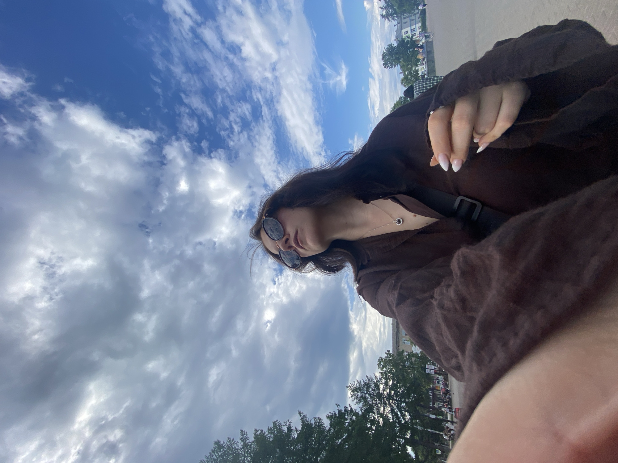 The image depicts a young woman with long, dark hair and sunglasses, looking up at a cloudy sky. She is wearing a brown shirt and a necklace with a circular pendant. Her right hand, with manicured fingernails, is visible in the foreground. The background shows trees, buildings, and other people in what appears to be a public outdoor space, possibly a park or a square. The sky is a mix of blue and grey clouds. The overall impression is of a casual moment captured outdoors, likely in the city of Chaikovsky, Russia, given the contextual information.