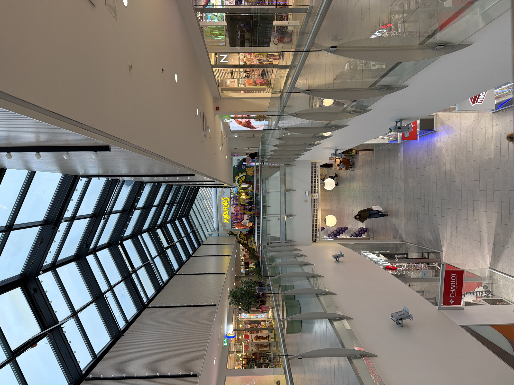 The image shows an interior view of a modern shopping mall, likely located in Tallinn, Estonia, given the presence of the Estonian language on signage. The mall features a multi-level design with escalators and walkways. A prominent glass and metal roof structure allows natural light to filter into the space. On the upper level, there is a colorful play area with a sign that reads "Merimors." Various shops are visible on both levels, including "Charlot," indicated by red signage, and "N" which appears to be a clothing store. People are seen walking and browsing in the mall. The environment is clean and well-lit, with a contemporary architectural style.