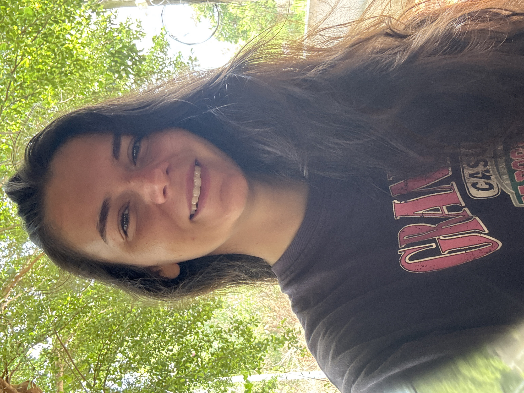 A woman with long, dark, wavy hair smiles at the camera. She is wearing a dark t-shirt with a faded red and white graphic that says "GRAND" in large letters. The woman has light skin and appears to be outdoors, with lush green foliage and dappled sunlight in the background, suggesting a tropical or subtropical environment. The scene is captured from the chest up. The location is not specified in the image or crops.