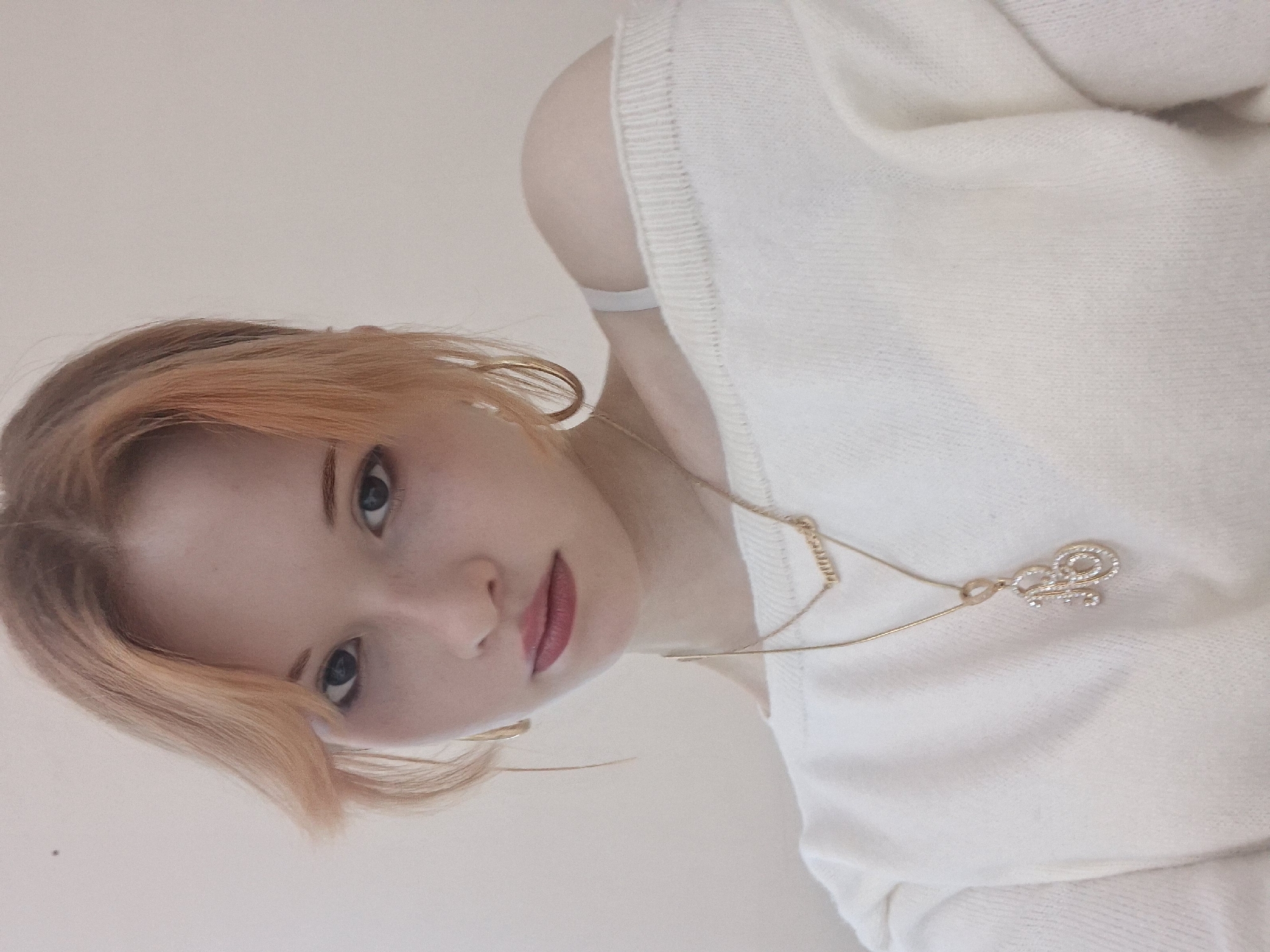 A close-up, rotated portrait captures a young individual, likely a selfie, in an indoor setting in Chaikovsky, Russia. The subject has fair skin, light-colored hair with peachy or light orange tones, and is looking directly at the camera with a neutral to slightly serious expression. Dark eyes and reddish-pink lipstick are prominent, along with subtle freckles on the nose and cheeks.
The person is wearing a light-colored, possibly off-white or cream, knitted sweater styled off-the-shoulder, revealing a bare shoulder and a light-colored bra strap. Gold-colored hoop earrings are visible on both ears. A delicate gold chain necklace drapes across the chest, featuring a sparkling pendant that appears to spell out the word "love" in a stylized script, adorned with small clear stones.
The background is a plain, light-colored surface, suggesting a simple wall or interior. The lighting is soft and even, highlighting the subject's features. The image is significantly rotated counter-clockwise, positioning the subject's head towards the bottom left of the frame, offering an unusual and dynamic perspective.