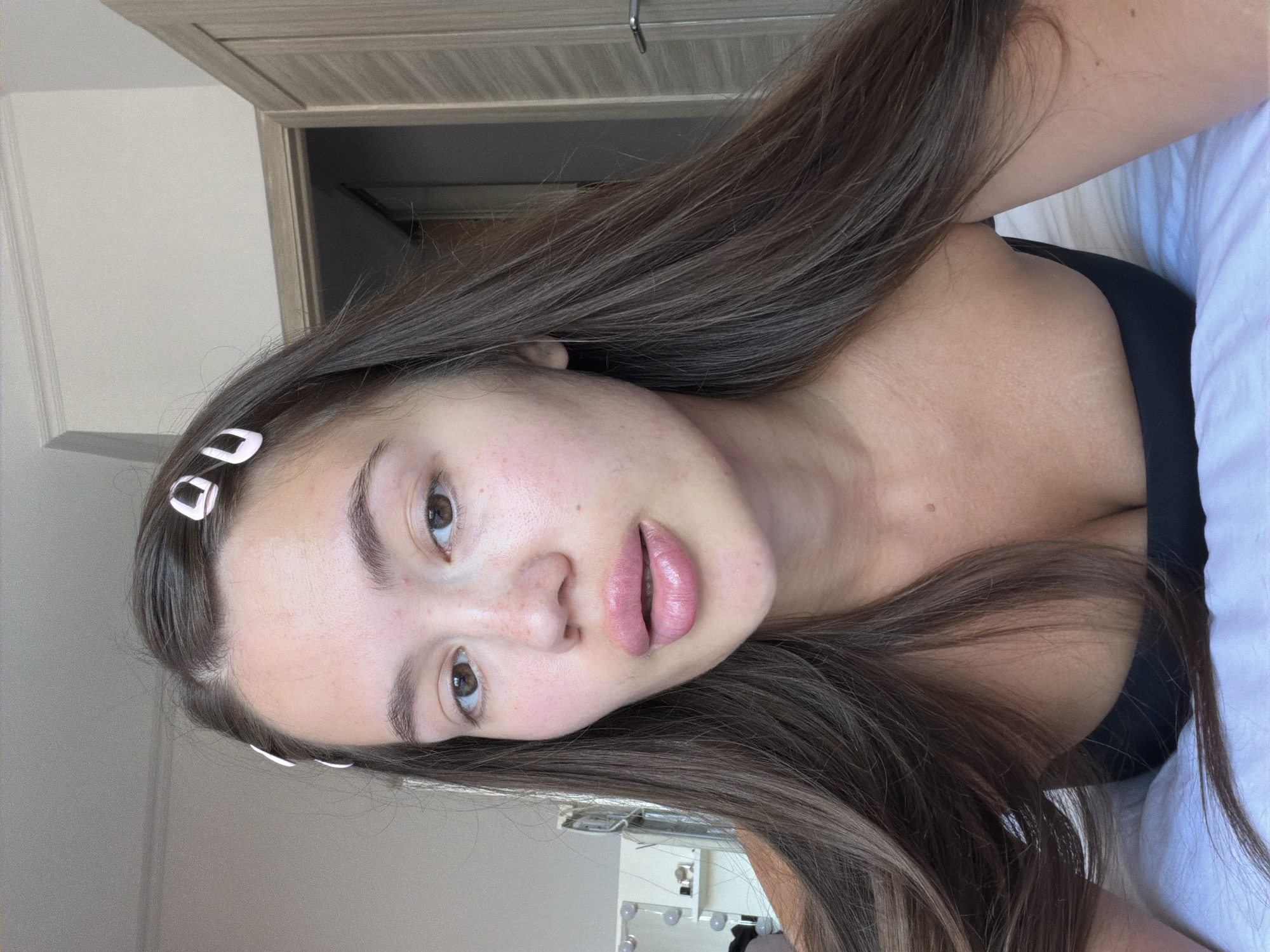 This is a close-up, selfie-style portrait of a young woman with long, brown hair and brown eyes. She is wearing a black top and has several small, pink hair clips in her hair. The background appears to be an indoor setting, possibly a bedroom, with a doorway and part of a door visible on the right. The lighting suggests it is daytime. There is no visible indication of the location being in Sirius, Russia.