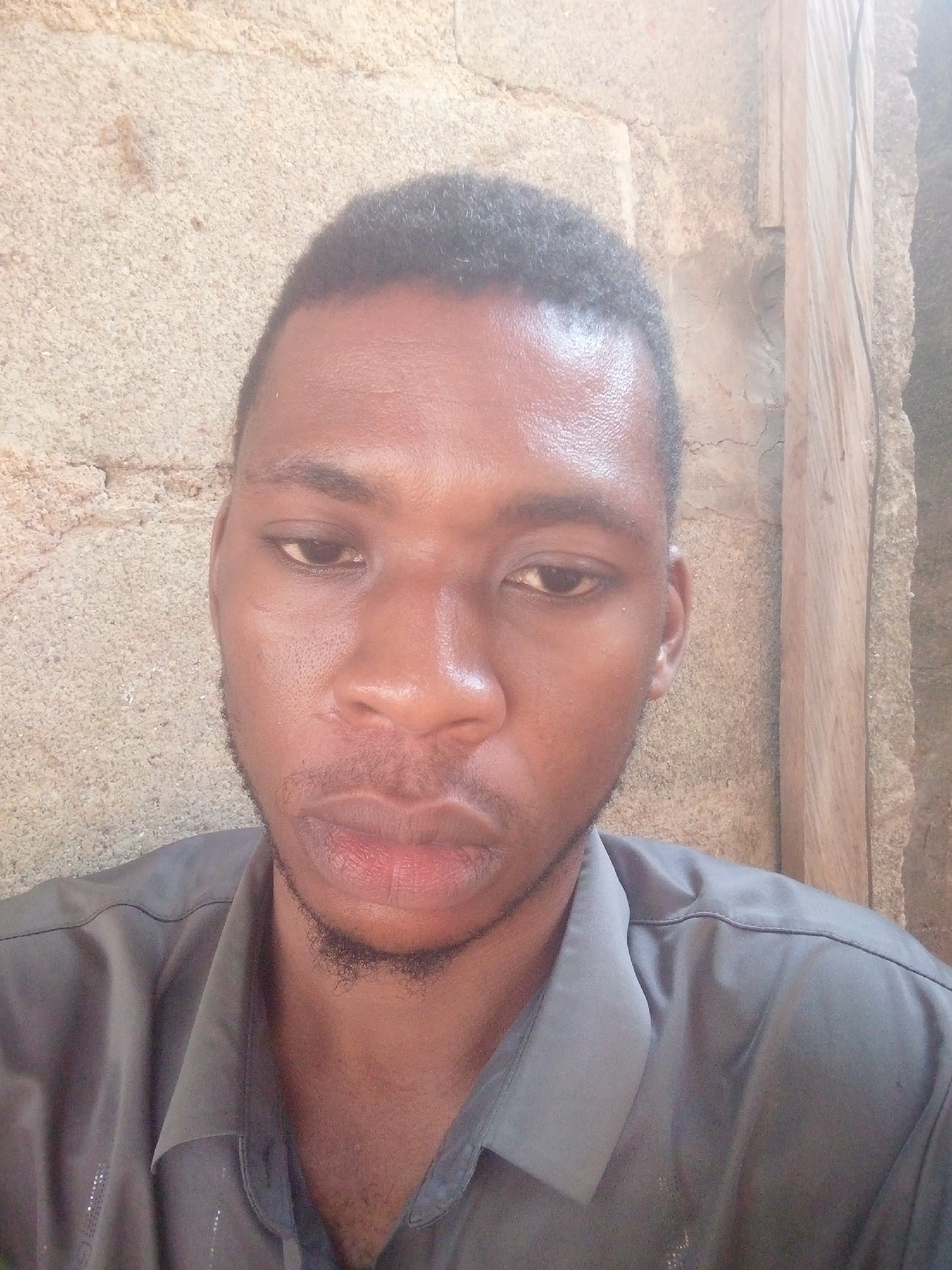 A close-up image shows a young man of apparent Black or African descent, likely a selfie. He has short, dark, textured hair and some faint facial hair, forming a goatee-like patch on his chin. His skin has a natural sheen, particularly noticeable on his forehead and nose. He is wearing a dark grey collared shirt, which appears to have subtle textured patterns or small reflective elements on the left side of the chest.
The man is looking downwards and slightly to his left, with a neutral, contemplative, or relaxed expression. His eyes are dark and his lips are full. The lighting suggests it is daytime, casting soft illumination on his face.
The background primarily consists of a light brown or beige textured wall, possibly made of stucco or rough concrete. On the right side of the image, a vertical wooden beam or frame is visible, set against the textured wall. The overall setting appears informal, possibly outdoors or in a simply finished indoor space. There is no visible text in the image.