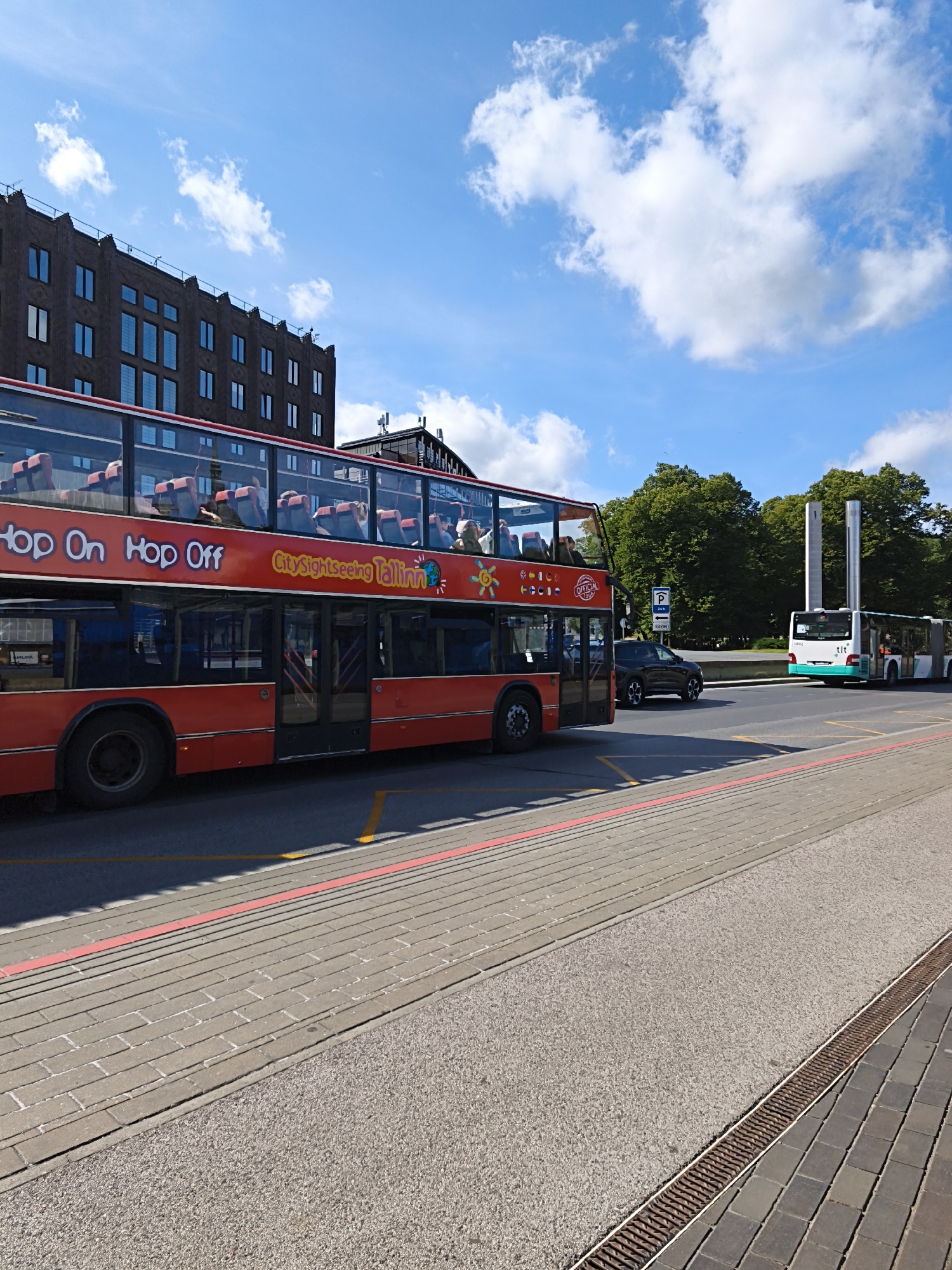 A bright, sunny day is depicted in Tallinn, Estonia, with a clear blue sky dotted with white cumulus clouds. The scene focuses on a red, double-decker "Hop On Hop Off" tour bus driving on a city street. The bus, labeled "City sightseeing Tallinn," has passengers visible on its upper deck. Next to the bus, a dark-colored car is also in motion. Further down the street, a white bus with green trim is driving, possibly a public transport vehicle. In the background, a tall, modern building with many windows stands, juxtaposed with lush green trees and a futuristic-looking metal sculpture or monument. The street is paved with brick and asphalt, with visible yellow and red painted lines demarcating traffic lanes and sidewalks. A metal drain grate runs along the edge of the pavement in the foreground. The overall impression is of a lively, urban environment on a pleasant day.