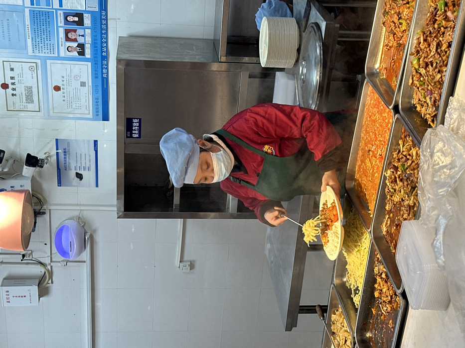 The image captures an indoor food service line within a cafeteria or canteen, identified as being in Jiangjin District, Chongqing City, China, by visible framed licenses on the wall. A female employee, positioned centrally, is depicted actively serving food. She wears a red long-sleeved jacket, a dark green apron, a light blue chef's hat, and a light blue surgical mask covering her nose and mouth. With a metal serving spoon, she is transferring yellow noodles from a stainless steel tray onto a white, multi-compartment plate she holds in her left hand. The plate already contains white rice and a reddish-brown saucy dish.
Numerous other stainless steel trays containing various hot food items, including different noodle preparations, saucy meat and vegetable dishes, are arranged along a counter in front of the employee, with steam visibly rising from some. To the right, a stack of clean, white multi-compartment plates rests on a stainless steel surface next to a covered pot. A tall, two-door stainless steel cabinet labeled "餐具存放柜" (Cutlery Storage Cabinet) is situated behind the employee.
The background consists of white tiled walls. Mounted on the wall are various items: security cameras (CCTV), a white wall-mounted air purifier emitting a blue light, and multiple framed documents including a "营业执照" (Business License), a "食品经营许可证" (Food Business License), and a poster describing an "AI 行为识别摄像机" (AI Behavior Recognition Camera). An overhead peach-colored light fixture illuminates the scene.