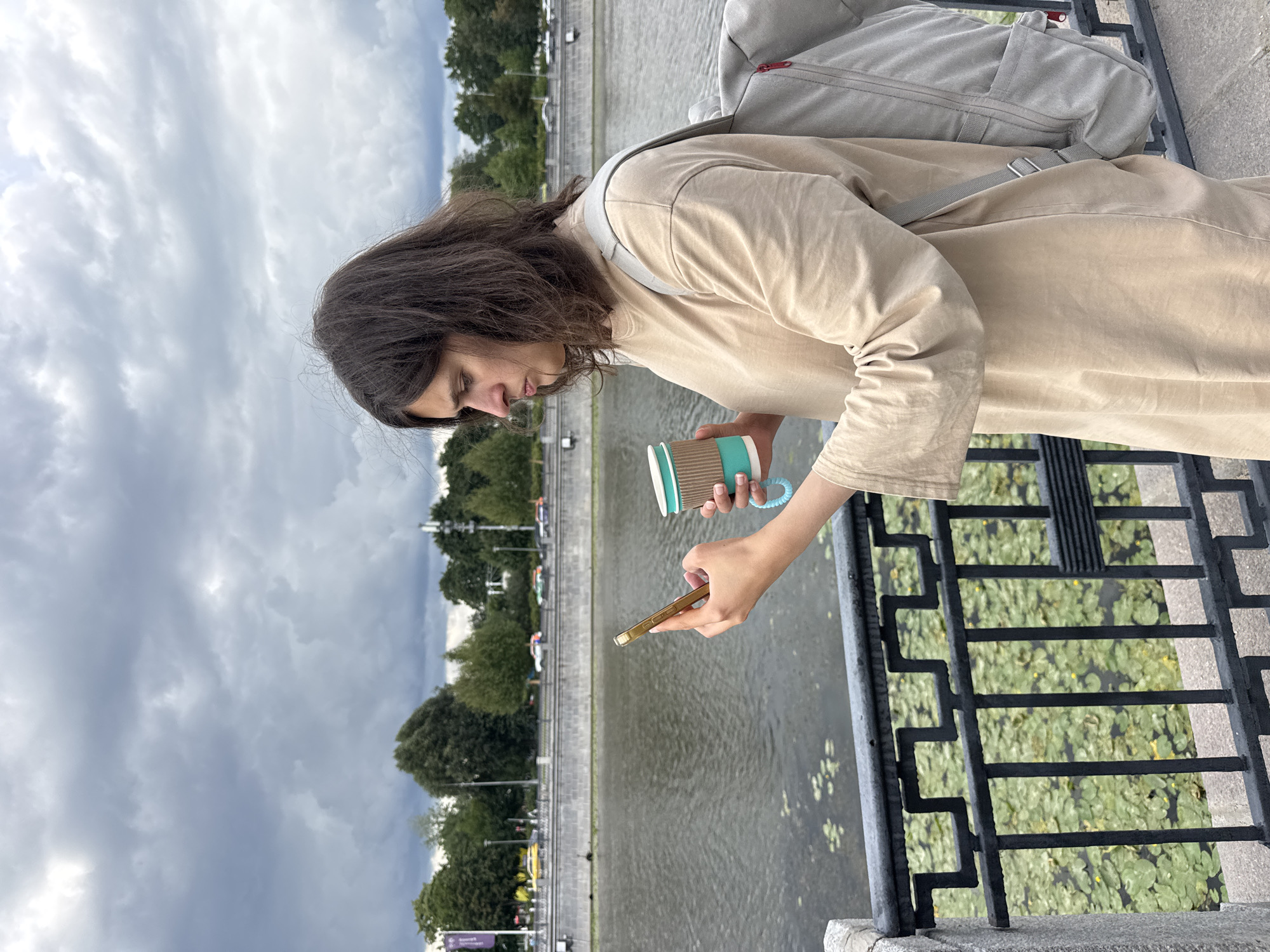 A young woman with dark, shoulder-length hair stands on a walkway next to a body of water, holding a smartphone in her right hand and a disposable coffee cup in her left. She is wearing a light beige, long-sleeved tunic and a light gray backpack is slung over her right shoulder. The backpack has red zipper pulls. The walkway appears to be bordered by a black metal railing with an ornate pattern. The water behind the railing is covered with lily pads and has a few small yellow flowers visible. In the background, across the water, is a tree-lined embankment with a road, cars, and buildings. The sky is overcast with dark, heavy clouds. The scene is likely in Moscow, Russia, given the architecture and vegetation. The woman is looking down at her phone, appearing to be engrossed in its use.