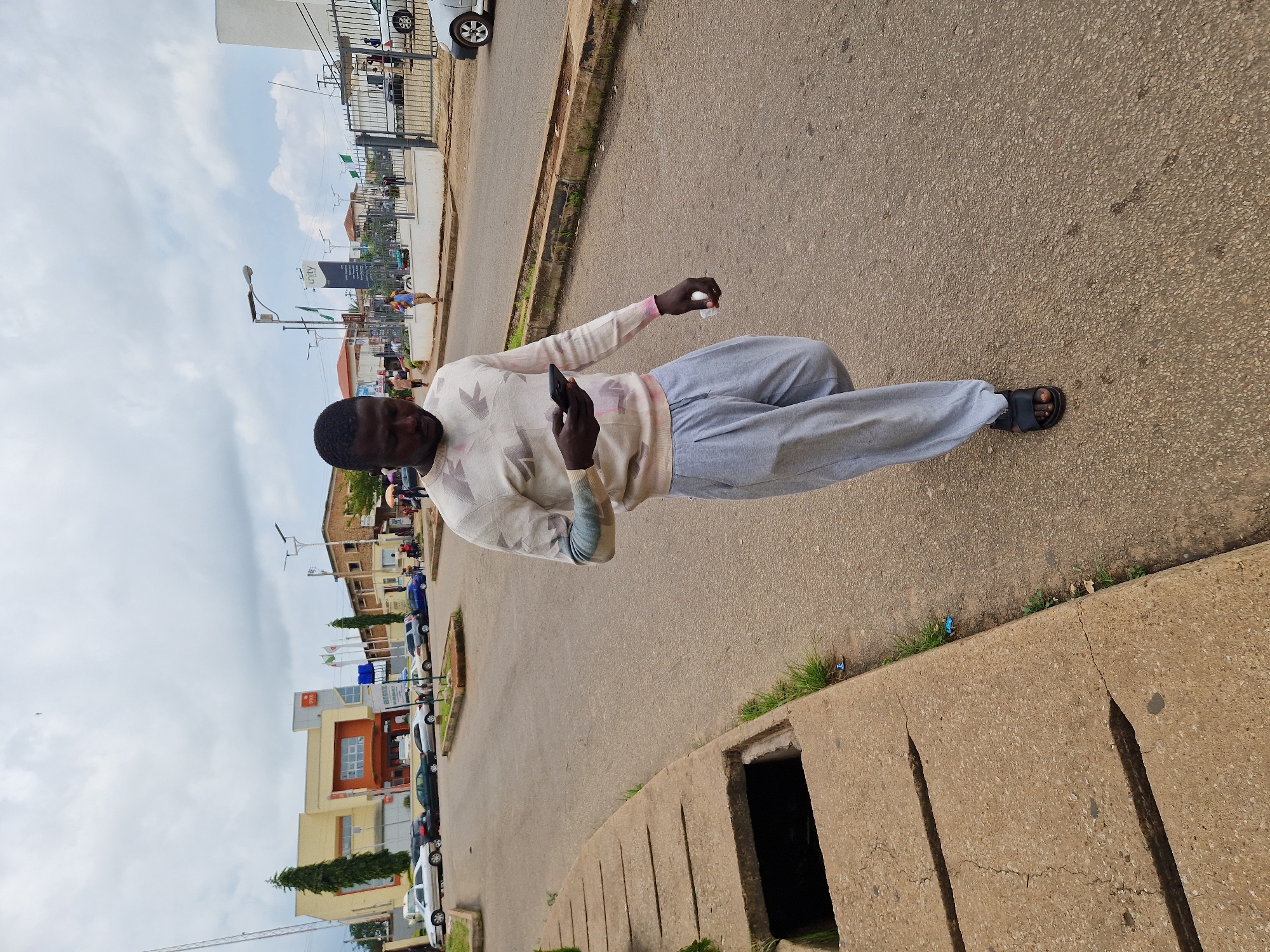 A man walks on a paved road in an urban environment, looking at his mobile phone. He is wearing a light-colored sweater with a geometric pattern and gray sweatpants. His feet are clad in dark sandals. To his left, a concrete drainage channel runs along the side of the road. In the background, buildings and parked cars are visible, under a cloudy sky.