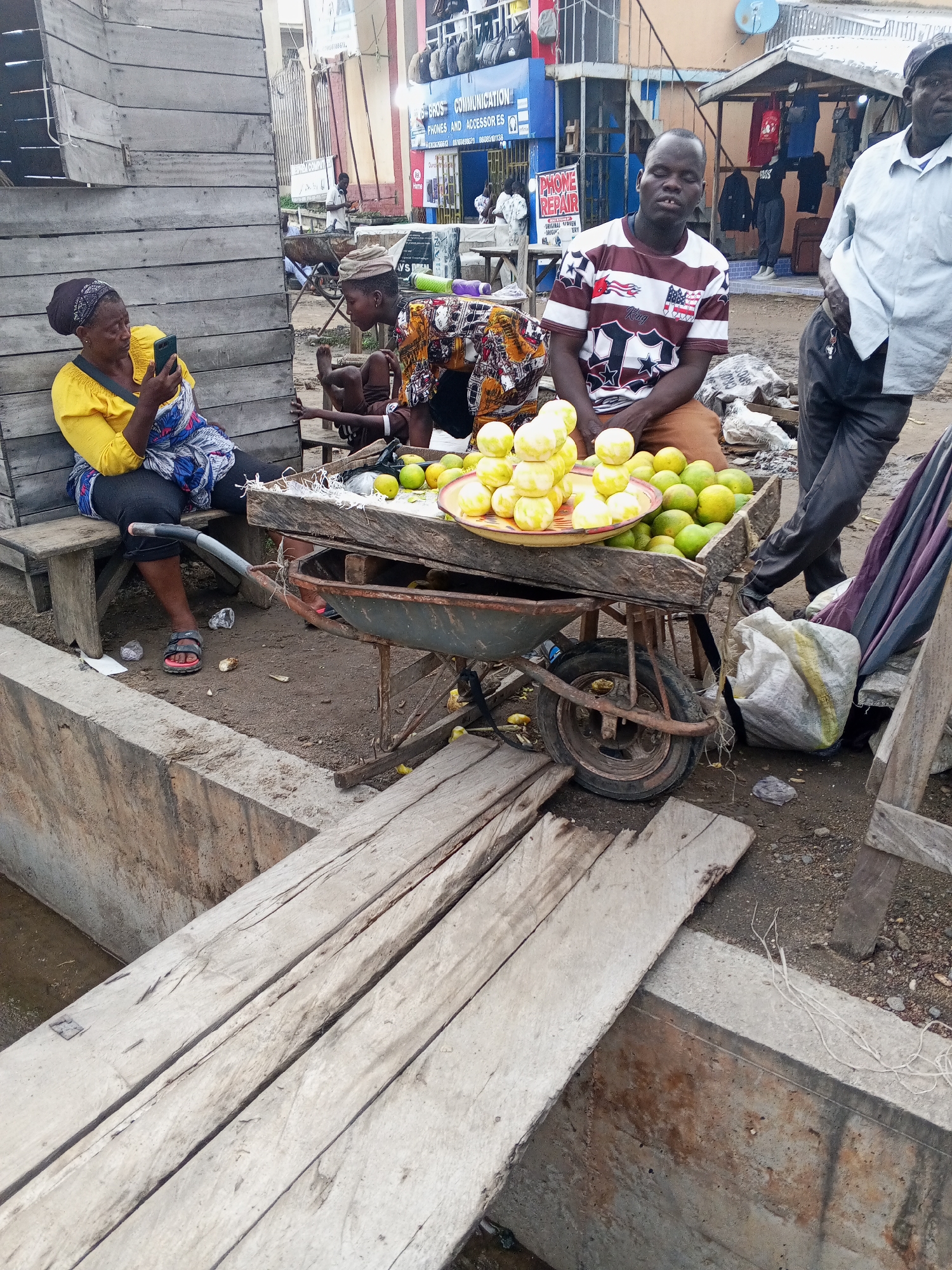 In Lokoja, Nigeria, a woman in a yellow top and a blue and white patterned skirt sits on a wooden bench, looking at her smartphone. Beside her, a young person in a colorful patterned shirt and a cap is also seated. In the foreground, a wooden cart with two wheels is laden with a pile of yellow fruits, likely lemons or limes, and a larger quantity of green fruits. Behind the cart, a man in a striped t-shirt and brown pants is visible, looking towards the camera with a neutral expression. To the right, another man in a light blue shirt and dark pants stands, with his right hand in his pocket. The scene appears to be an outdoor market or roadside stall. In the background, there are buildings with signs indicating businesses, one of which reads "G-BROS COMMUNICATION PHONES AND ACCESSORIES". A wooden plank walkway is visible in the lower part of the image, leading over a shallow, water-filled channel. The ground is covered with dirt and gravel.