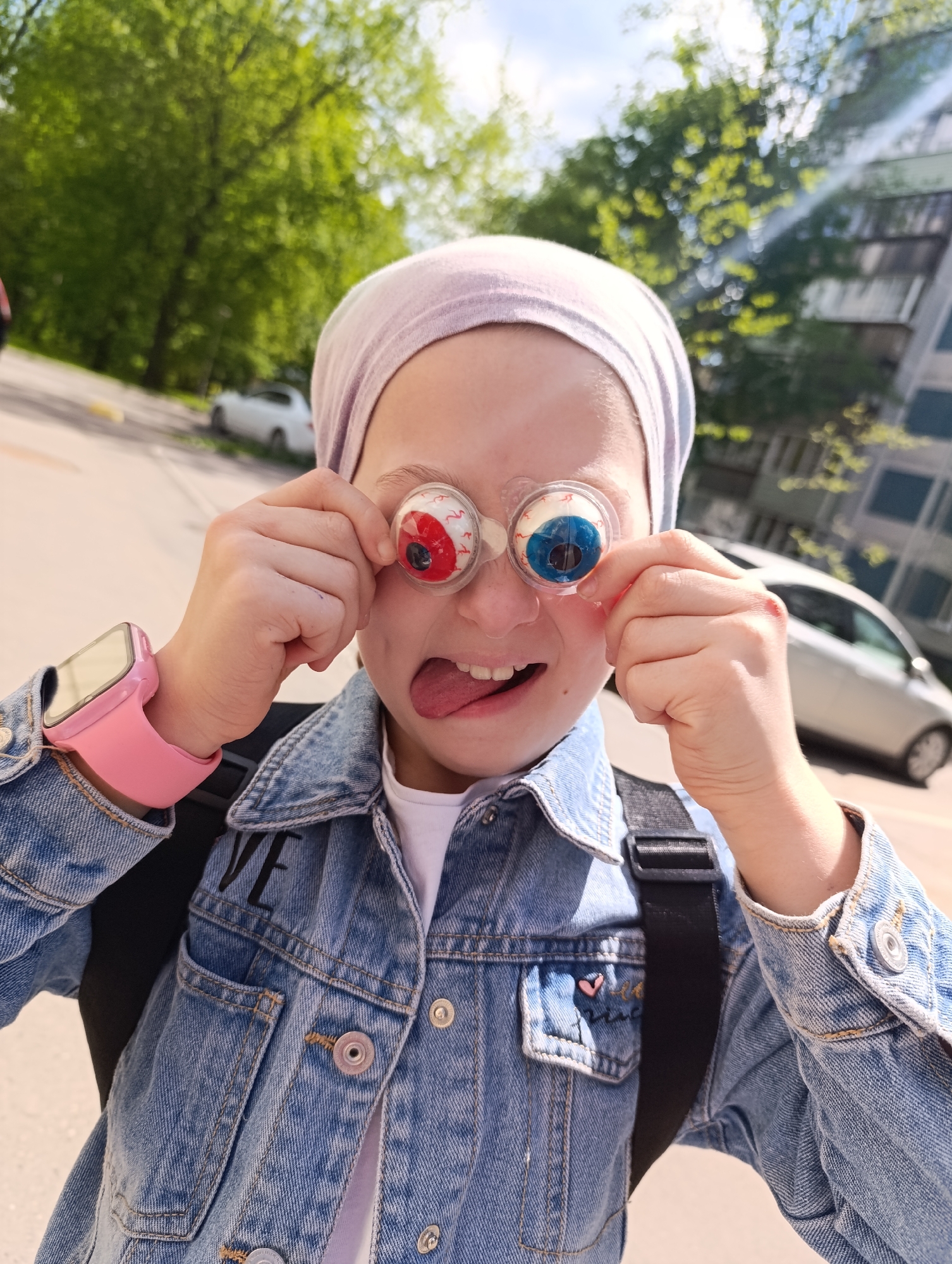 A girl is standing outdoors in an urban environment. She is wearing a light purple head covering, a denim jacket, and a pink smartwatch on her left wrist. She is holding novelty eyeball glasses up to her face, sticking her tongue out, and making a silly face. Behind her are parked cars, a building, and trees. The city and region are unknown.