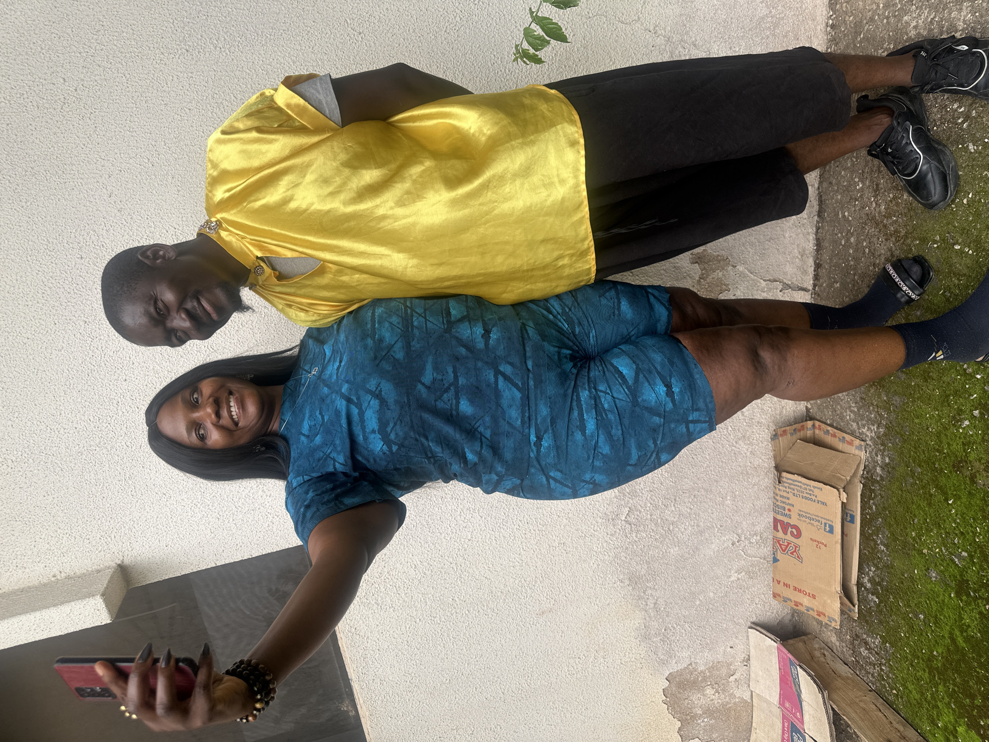A Nigerian man and woman pose for a selfie outdoors in front of a textured white wall. The woman, on the left, is wearing a blue and black patterned t-shirt and shorts set, and is holding a red-cased mobile phone to take the photo. She has dark hair and is smiling. The man, on the right, is wearing a bright yellow top with a grey t-shirt underneath and black shorts. He has short, dark hair and a goatee, and is looking away from the camera. In the background, the wall is weathered and there are some cardboard boxes and mossy ground visible at the bottom of the frame, suggesting an outdoor urban environment, possibly a residential area in Jos, Nigeria.