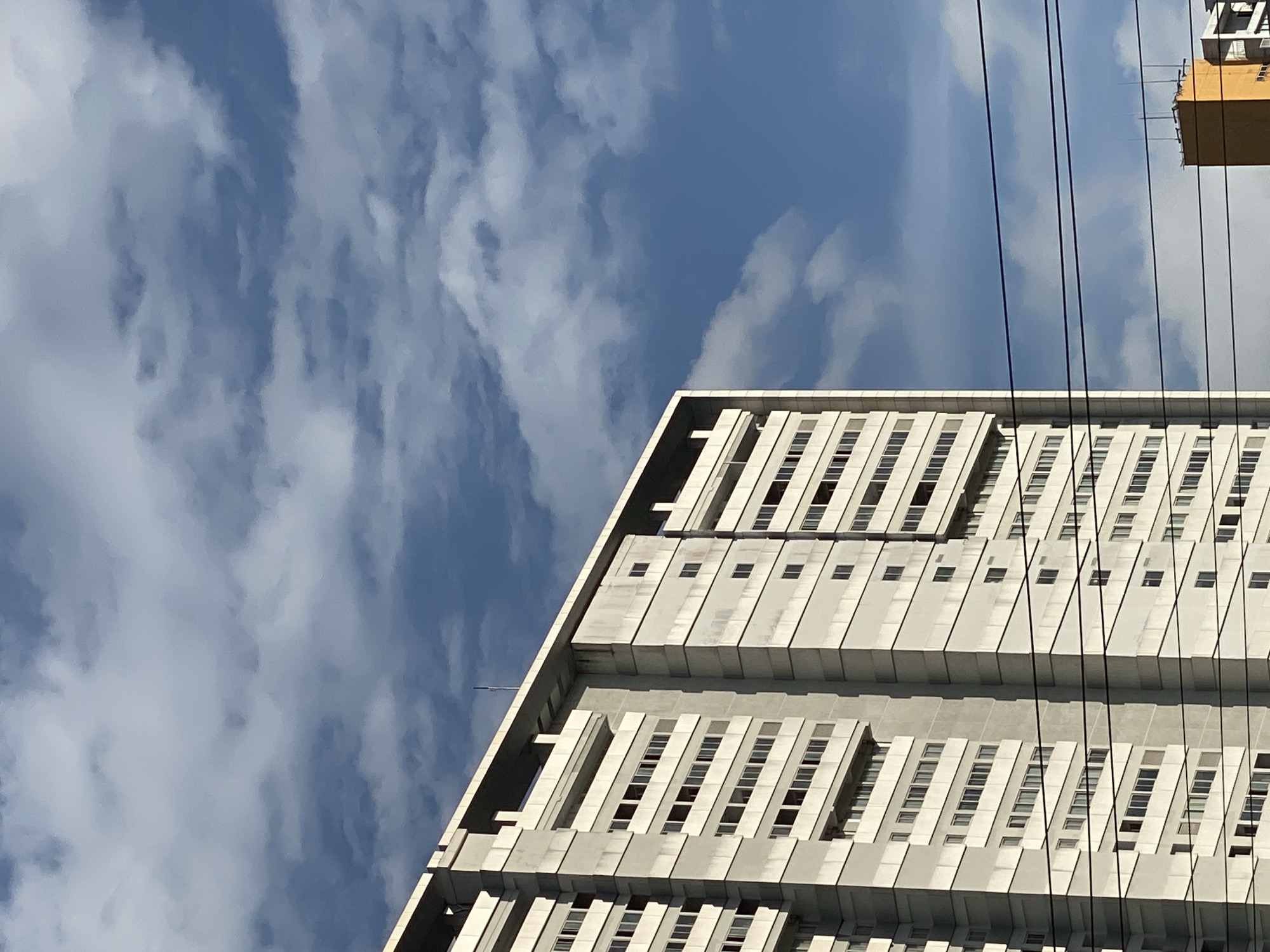 A medium shot captures the side of a tall, light-colored apartment building against a bright blue sky with wispy white clouds. The building has a linear design with repetitive rows of windows. Dark horizontal wires are visible across the lower portion of the frame, obscuring some of the building's lower levels and a small, orange building peeking out from the far right. The image appears to be taken in Gambut Barat, Indonesia, on a clear day. There are no people or animals visible in the frame.