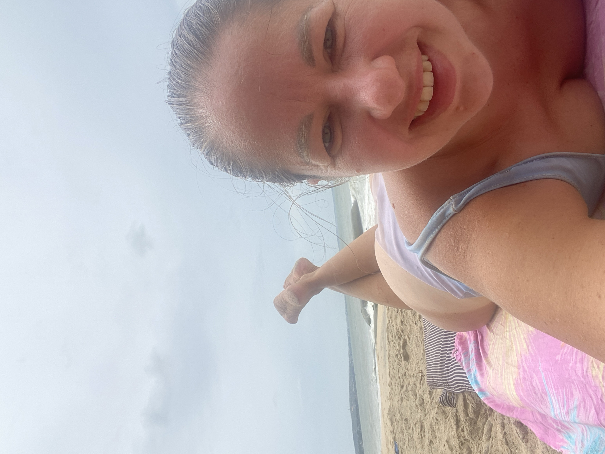 A woman is lying on a beach towel on the sand, facing the camera. She is wearing a light blue bikini. Her legs are crossed and her feet are in the air, showing they are covered in sand. The ocean and a cloudy sky are visible in the background. The woman is smiling broadly.