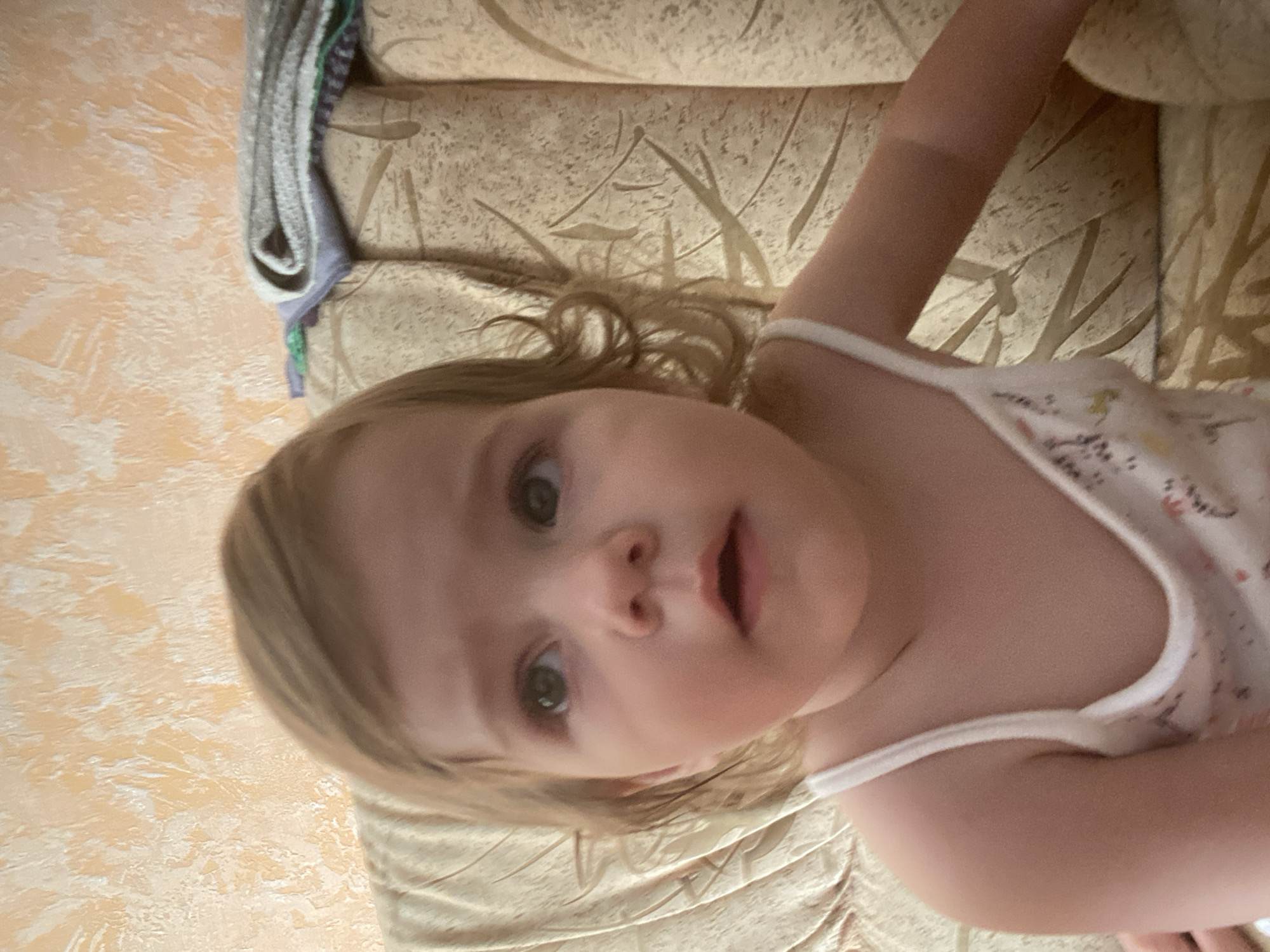 An overhead medium shot shows a young Caucasian child with light brown hair, green eyes, and fair skin sitting on a couch. The child is looking up and to the left with a neutral expression. The child is wearing a white tank top with a floral pattern. The couch is beige with a textured pattern. In the background, a wall with a textured orange and white pattern is visible. A folded blanket with gray and purple stripes is on the back of the couch.