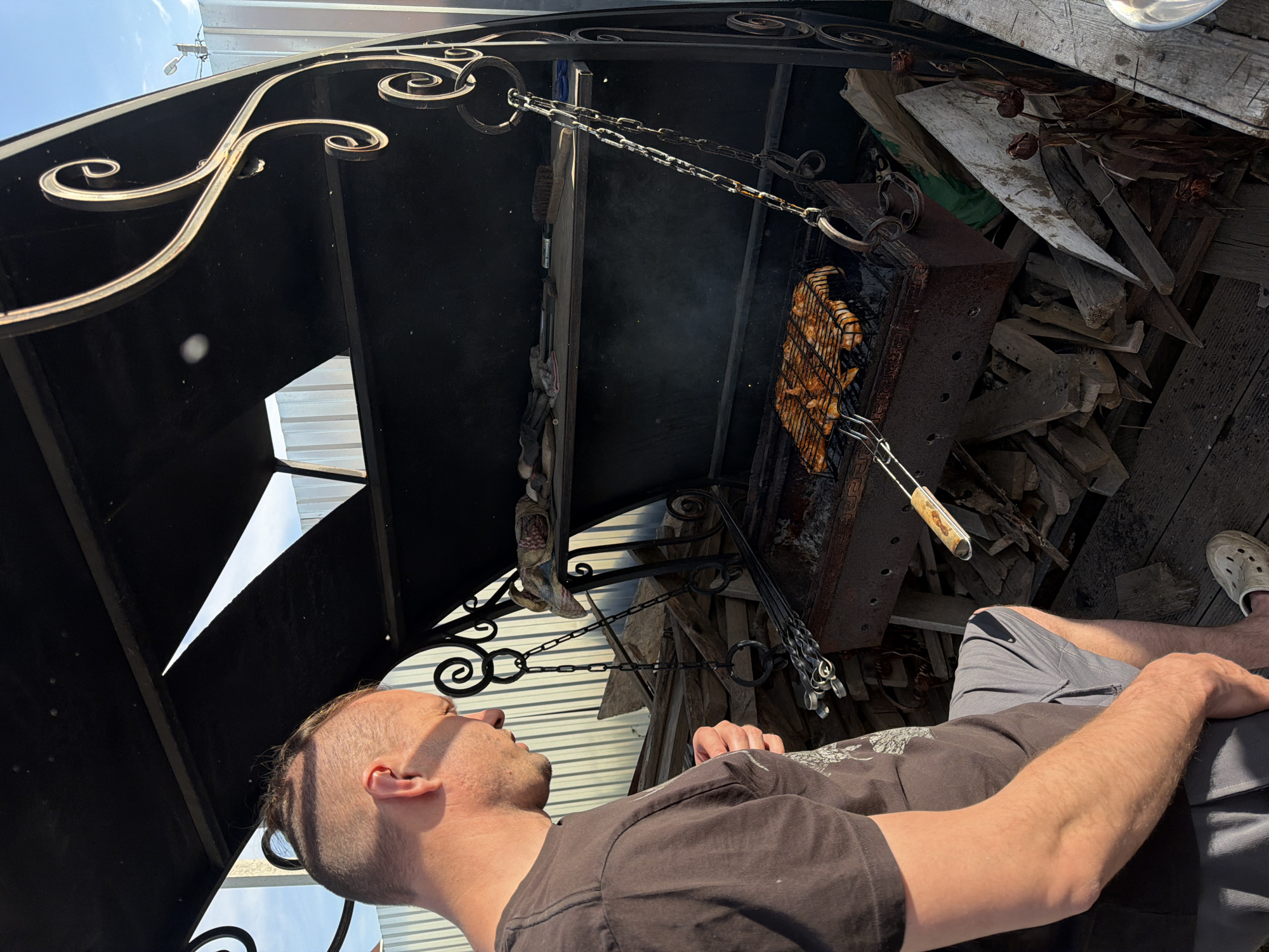 The scene depicts a man in Chaikovsky, Russia, grilling food. He is wearing a dark t-shirt and grey shorts and is seated. A rusty metal grill with food inside hangs from a chain suspended by an ornate wrought iron structure. The grill appears to be over a fire pit with wood nearby. The floor is made of wooden planks.