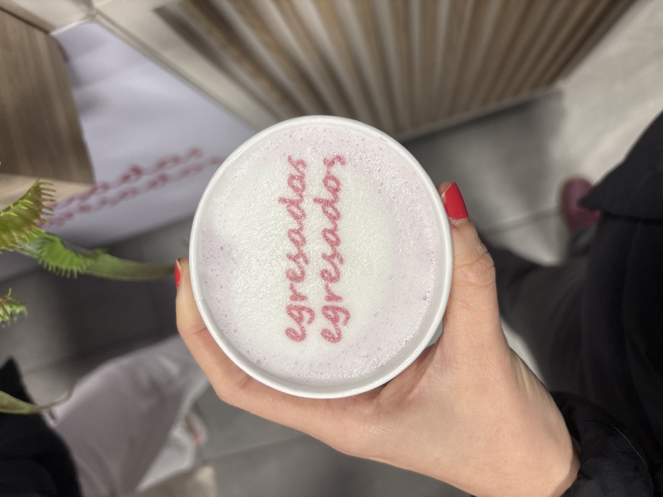The image captures a close-up, top-down view of a white disposable cup held by a human hand. The hand, featuring red manicured fingernails, grasps the cup from the right side. The cup's contents are topped with white and light purple foam, upon which pink, granulated sugar forms the words "egresadas" on the upper line and "egresados" on the lower line.
The individual holding the cup wears a dark long-sleeved garment. In the lower frame, dark trousers and a reddish-pink shoe are visible against a light-colored floor. The background is shallowly focused and blurred. A green plant with serrated leaves is partially visible in the upper background. To the upper left, an indistinct figure in light-colored clothing is present. To the right, a vertically-textured light-colored wall or panel is discernible.
The scene is indoors, consistent with a commercial establishment such as a café or an event space. The precise location within Mexico City, Mexico, is not discernible from the provided visual information.