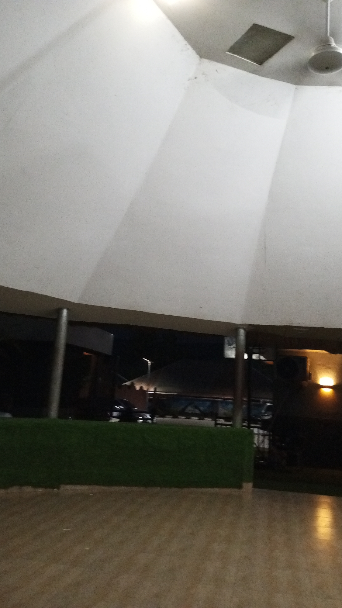 The image shows the interior of a covered space in Adankolo, Nigeria. The roof is white and domed, with a ceiling fan and a square vent visible. Below the roof, there are support pillars. A green barrier is visible. In the background, through the openings, there are structures and vehicles suggesting an outdoor area at night. The floor is tiled in a light brown pattern. There are no people visible in the frame. A light source casts a glow in the background.