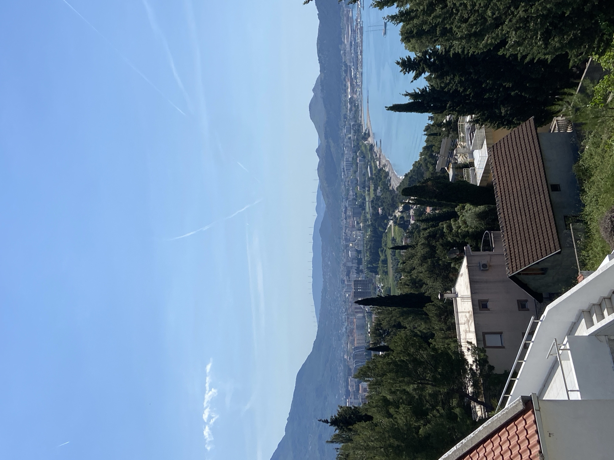 The image presents a panoramic view of a vibrant coastal town, identified as Šušanj, Montenegro, from an elevated perspective. The scene is bathed in bright daylight under a clear, azure sky, marked by several thin, white contrails from aircraft.
In the immediate foreground, on the right side of the frame, are parts of residential structures and dense greenery. A prominent white multi-story building with balconies and an external white staircase descends downwards, while another building with a reddish-brown tiled roof is partially visible. Tall, dark green coniferous trees frame the view, suggesting a lush environment.
The midground showcases the heart of the coastal settlement, stretching along a narrow strip between the sea and the mountains. A multitude of buildings, both residential and possibly commercial, line the shore. A long, slender beach curves along the coastline, meeting the tranquil, light blue waters of the bay or sea. Several boats are visible in the water, and a more developed port or harbor area can be discerned further down the coast.
The background is defined by majestic mountains that rise sharply from the water's edge. The mountains closer to the viewer are covered in verdant foliage, while those in the distance appear softened by atmospheric haze. Faintly visible along the crest of a distant ridge are several vertical structures, possibly wind turbines.
The image captures a serene and picturesque daytime scene, likely in the morning or early afternoon, given the clear sky and bright illumination. There are no people visible, nor is there any discernible text within the image.