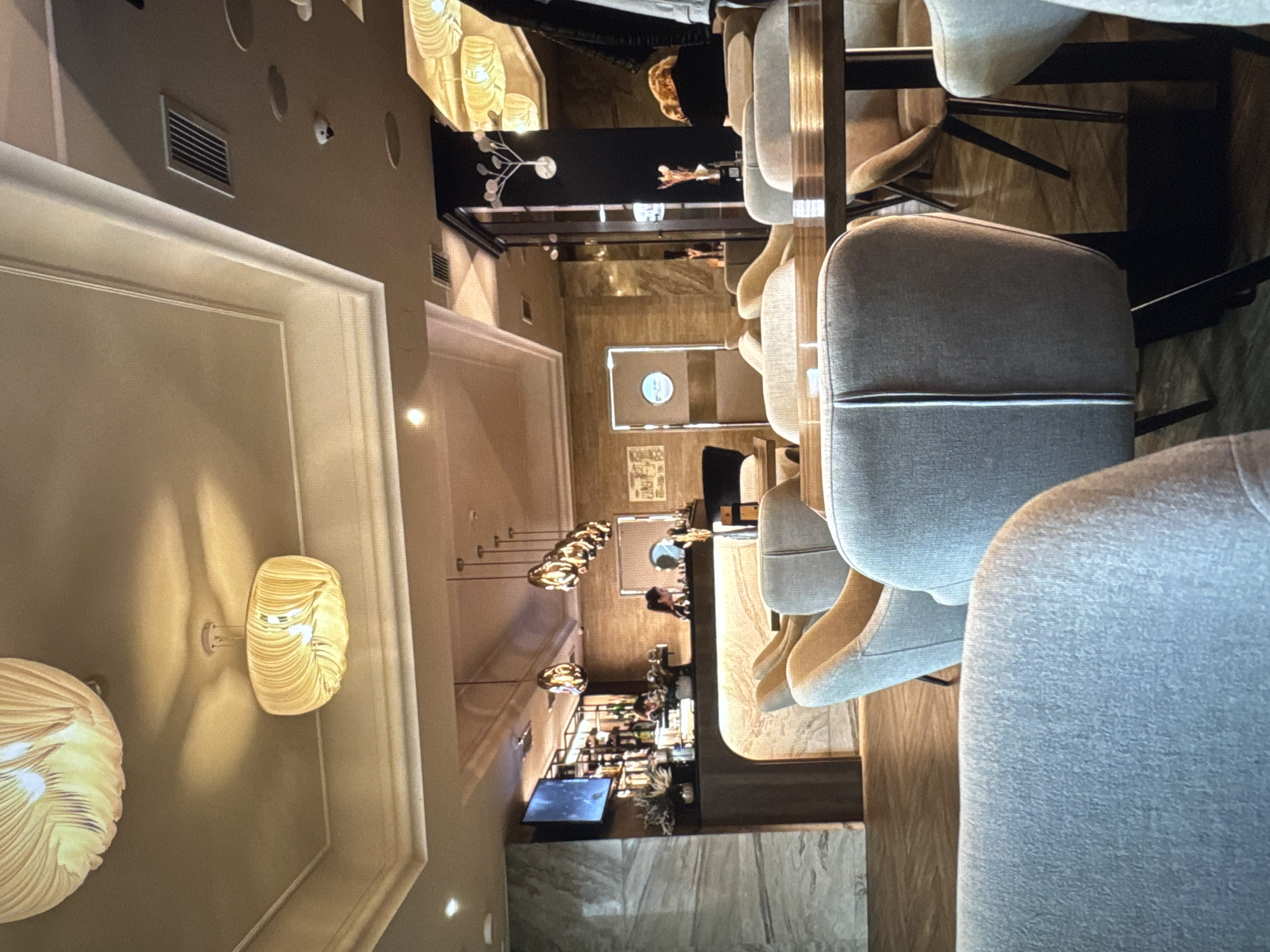 The image shows the interior of a modern cafe, possibly located in Novinki, Russia. The cafe features a well-lit, contemporary design with a bar area, seating arrangements, and decorative elements.
In the foreground, several plush, light-colored armchairs are visible, suggesting a comfortable dining or lounging experience. The chairs have a textured fabric and a modern, streamlined design. One chair in the center is particularly prominent, showcasing its cushioned backrest with a vertical seam detail.
The middle ground reveals a bar area with a marble-effect counter and a backlit surface. Behind the counter, shelves stocked with bottles and glasses are illuminated, indicating a bar setup. Two individuals appear to be working behind the bar, one of whom is partially visible. A television screen mounted on the wall displays an image.
The background includes a series of pendant lights with an intricate, textured design, casting a warm glow. The ceiling is adorned with molding, and ventilation grilles are visible. Further back, there are doors with circular signage and decorative elements on a dark pillar. The flooring appears to be a polished wooden surface, reflecting the ambient light. The overall ambiance is one of a sophisticated and inviting establishment.