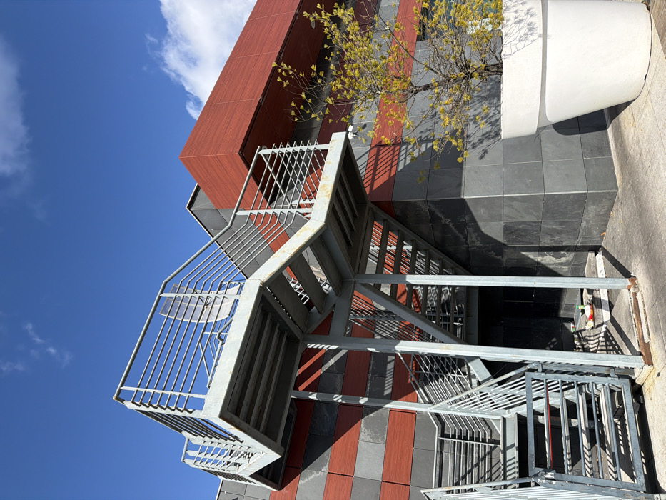 The image captures an exterior view of a modern building in an urban area of Viseu, Portugal, under a clear blue sky with scattered white clouds. A multi-flight external metal staircase, constructed from galvanized steel with horizontal bar railings and open risers, ascends against the building's facade on the left. The building's lower section is clad in dark grey horizontal panels, interspersed with narrower horizontal bands of reddish-orange material. An upper section of the building exhibits a larger expanse of reddish-orange cladding with sharp architectural lines. Direct sunlight casts distinct shadows from the staircase onto the building facade and the paved ground below. Beneath the lowest flight of stairs, near a dark recessed doorway, a white plastic bottle with a red cap and other small discarded items are visible on the ground. To the right of the staircase, a small, deciduous tree with sparse, nascent yellow-green leaves is planted in a large, white, conical planter pot. A white security camera is affixed to a reddish-orange band on the building facade, positioned above the tree. No individuals are present in the scene, and no overt actions or interactions are depicted.