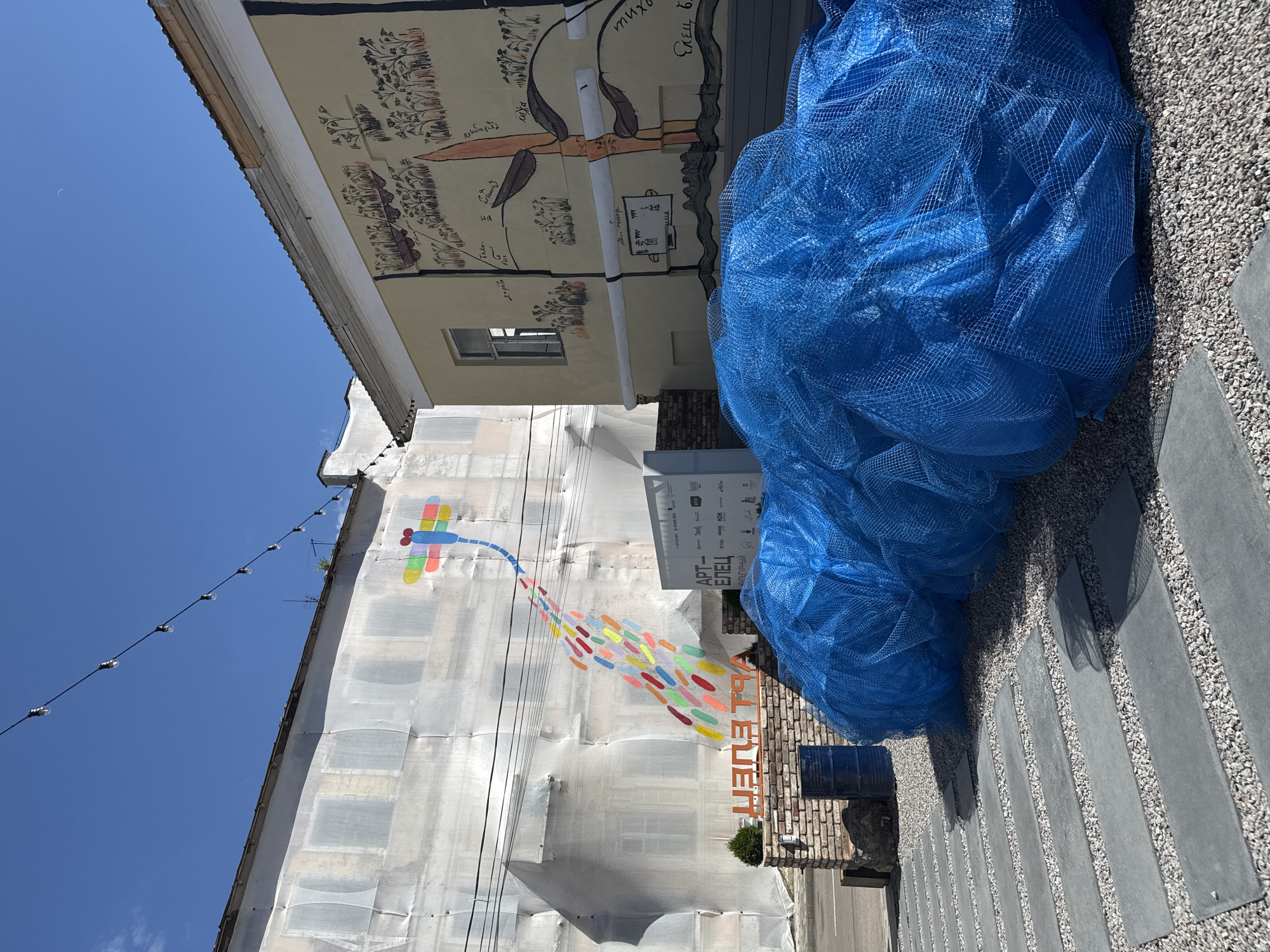 The image depicts an outdoor scene in the city of Yelets, Russia, likely a public space or courtyard. In the background, a building is covered in white protective sheeting, with a colorful mural of a dragonfly and a trail of colorful dots painted on it. To the right, another building features a mural with stylized drawings of plants and figures, rendered in shades of yellow and brown. A light string with bulbs stretches across the top of the scene. In the foreground, a pile of large blue netting lies on a gravel surface interspersed with rectangular paving stones. A brick structure with a sign that reads "АРТ ЕЛЕЦ" (ART YELETS) is visible to the left of the netting, accompanied by two blue barrels and a small evergreen plant. The sky is clear and blue, with a faint crescent moon visible. No people are present in the image.