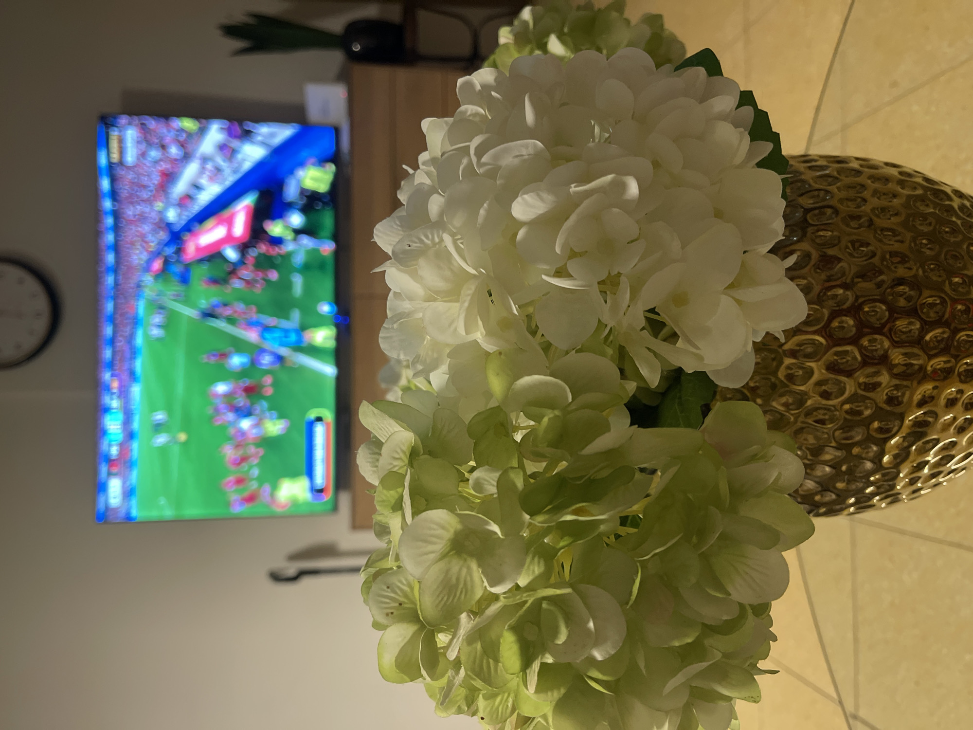 In the foreground, a close-up view shows a bouquet of white and pale green hydrangeas in a textured gold vase. The vase rests on a reflective glass surface, likely a table. In the background, a large television screen displays a vibrant, action-filled sports game, possibly soccer, with many spectators in the stands. The overall ambiance suggests a casual indoor setting, possibly a living room, with the television as a focal point. The lighting is subdued, creating a warm and intimate atmosphere. The scene is domestic and relaxed, with the flowers providing a decorative element to the entertainment. The location specified in Netanya, Israel is not visually evident in the provided image.