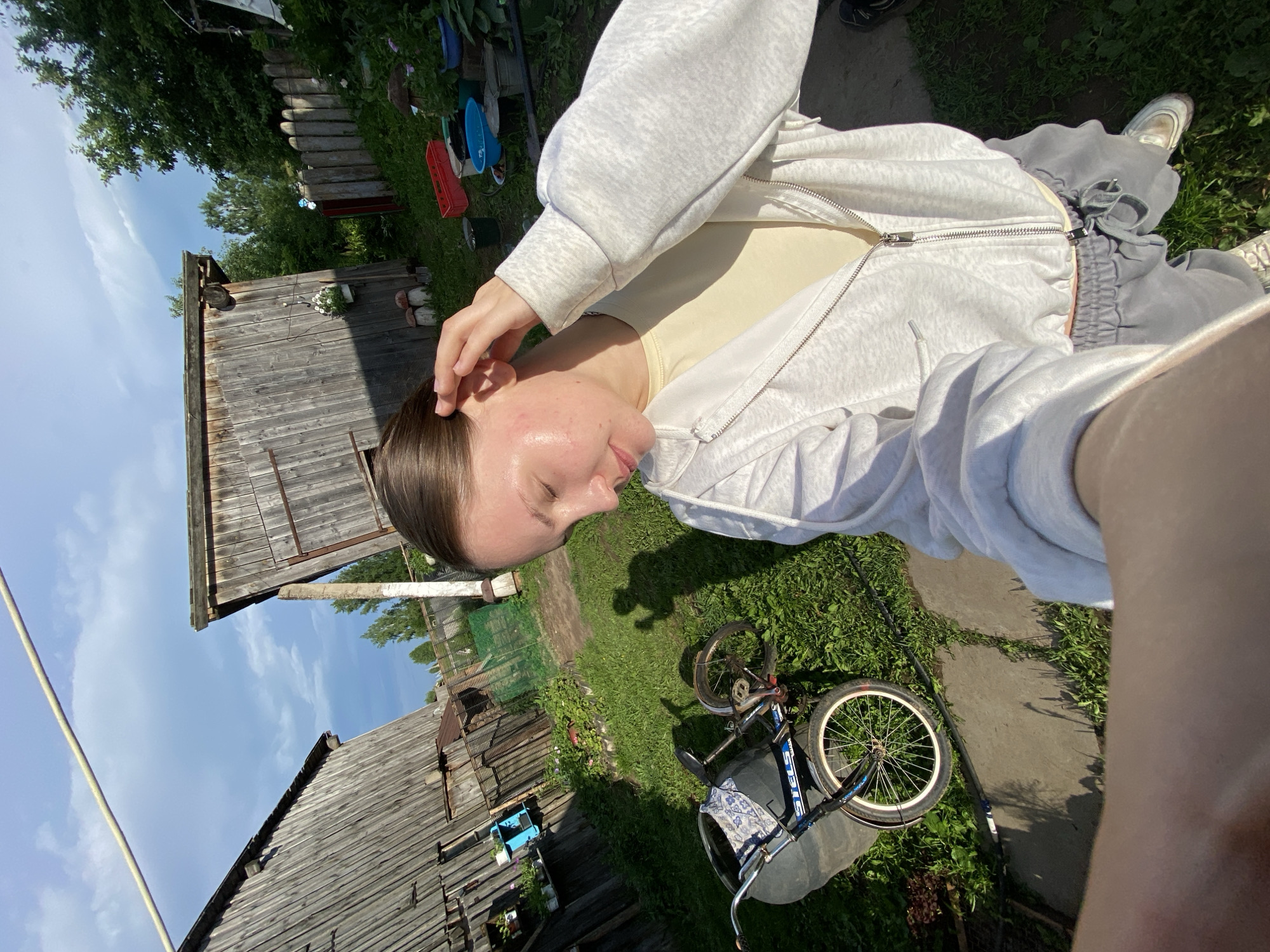 A young woman with brown hair pulled back is standing in a rural outdoor setting. She is wearing a light-colored hooded jacket over a yellow shirt and grey sweatpants. Her right hand is touching her ear. In the background, there are two weathered wooden structures that appear to be sheds or outbuildings. A bicycle with a basket is leaning on its side on the grass to her left. The ground is a mix of grass and dirt, with a concrete path visible. Various gardening items, including red and blue tubs, and buckets, are scattered around. Trees and a wooden fence are visible in the distance. The sky is blue with scattered clouds, and the sun is casting shadows, indicating it is daytime. The scene depicts a casual moment in a rural Russian village, likely in Вассята, given the context provided.