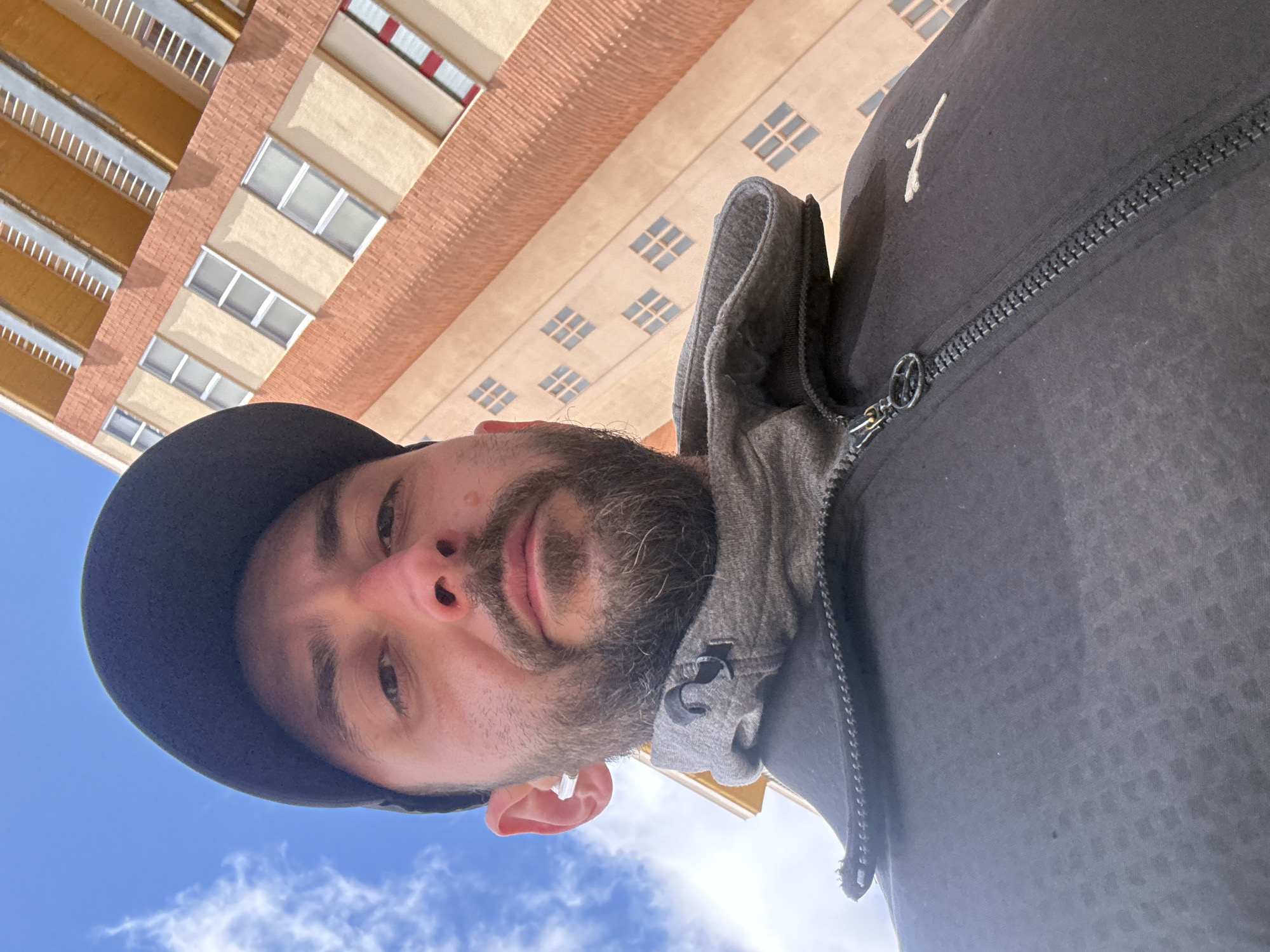 A selfie captures a bearded man wearing a black cap and dark gray hoodie standing beneath a building. The building, partially visible in the background, is several stories tall with a light-colored facade and brick details. Balconies with yellow railings line one side of the building. The sky, a clear blue with scattered clouds, is also visible in the upper left corner of the image. The location is Soria, Spain.